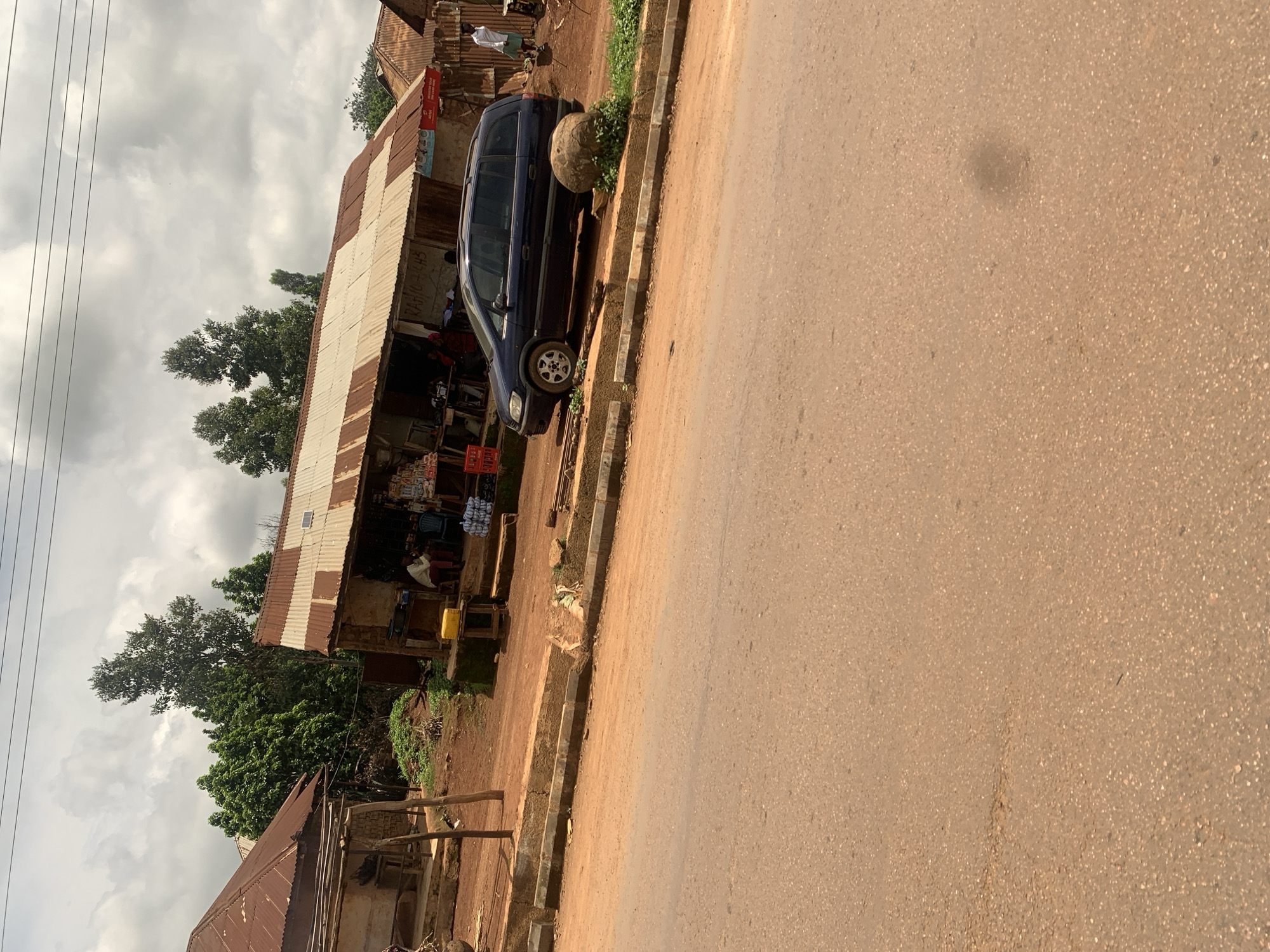 The image captures a street scene in Hoss, Nigeria, with a focus on a small roadside market. A dark blue minivan is parked in front of a weathered structure with a corrugated metal roof. The structure appears to be a shop or stall, with goods displayed along the front and a few people gathered inside and in front. A large, round rock sits beside the minivan. To the left, another building with a similar corrugated roof is visible. The ground is composed of red earth and gravel. The sky is overcast with clouds. The perspective is from across a paved road, which occupies the foreground.