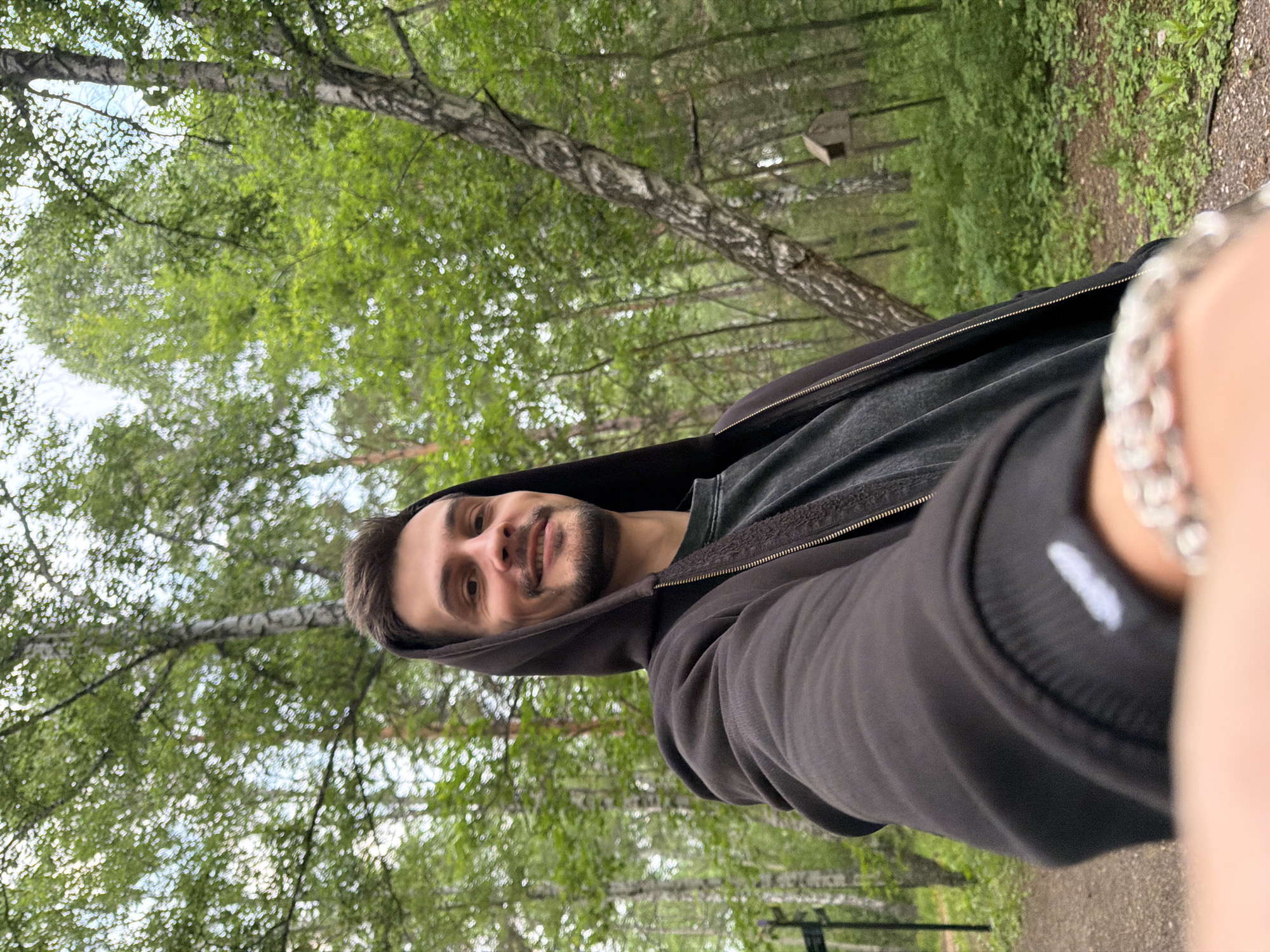 A selfie is taken by a young Caucasian man with short brown hair and a beard, smiling. He is wearing a dark hooded jacket zipped over a dark gray t-shirt. He has a silver bracelet on his left wrist, which is partially visible. The man is in a park with numerous birch trees, evident by their white bark and leafy green canopies. Lush green undergrowth and a dirt path are visible in the background. A small wooden birdhouse is mounted on a pole to the right of the frame. The lighting suggests it is daytime, possibly overcast. The location appears to be a natural area within or on the outskirts of Krasnoyarsk, Russia, given the prevalence of birch trees often found in Siberian forests.