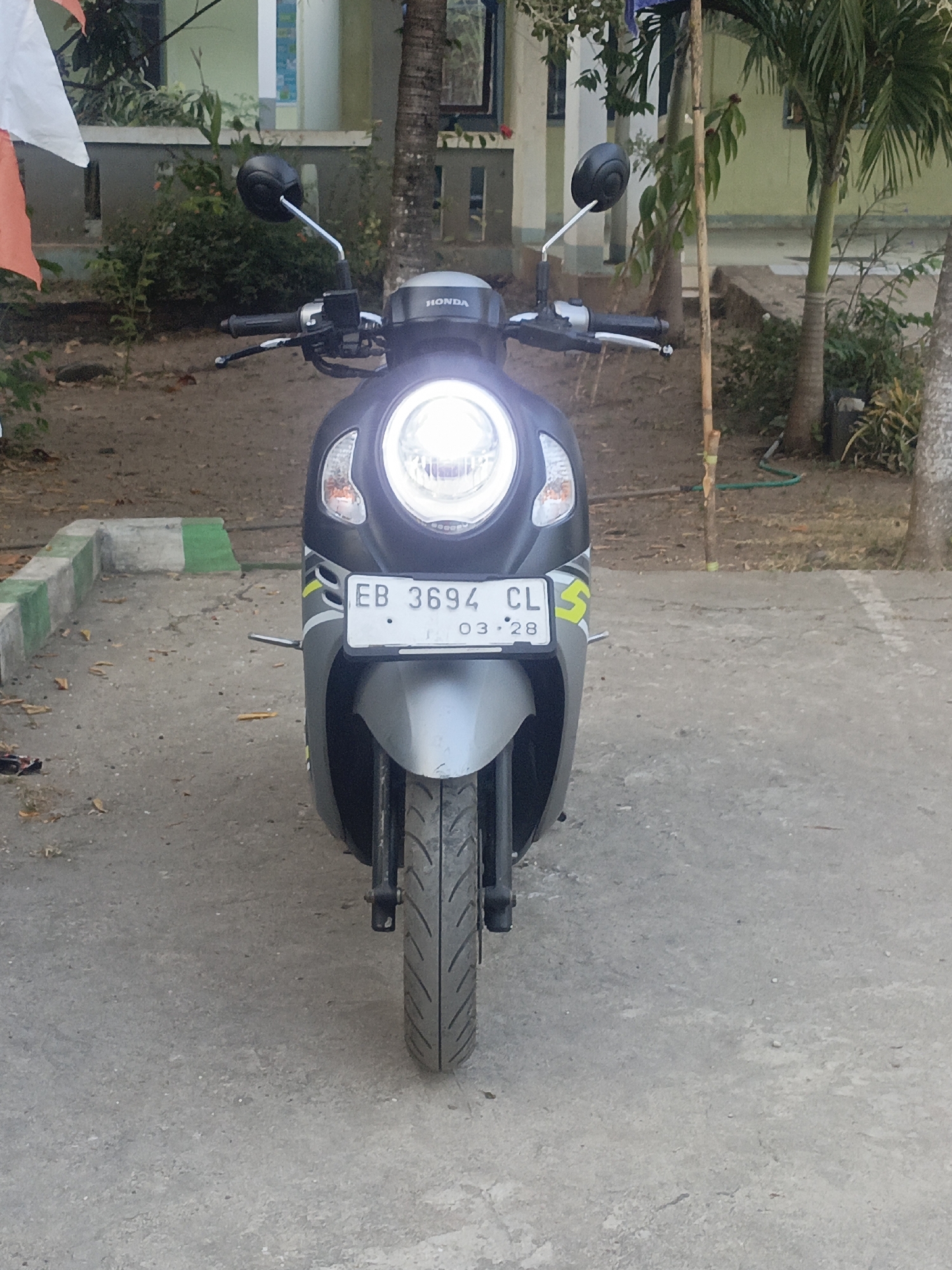 An eye-level, medium shot captures a parked, dark gray Honda scooter in what appears to be a residential or institutional courtyard in Solor Barat, Indonesia. The scooter's headlights are illuminated, casting a bright glow. Its license plate, positioned centrally, reads "EB 3694 CL" with "03 . 28" below it. The scooter is situated on a concrete surface, with a curb that is painted with alternating white and green sections. In the background, foliage from trees and shrubs is visible, along with parts of a building with pale green walls. A partial view of a white and orange flag is seen on the left side of the image, along with a rough wooden pole. The overall scene suggests a moment of stillness, with the scooter parked and unattended in an outdoor setting.