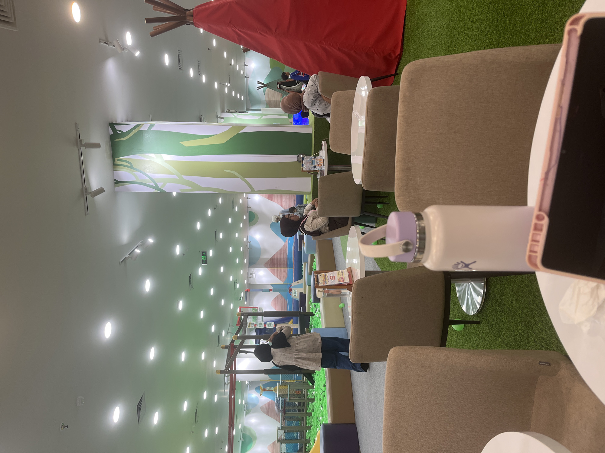 The image depicts an indoor children's play area with a cafe section. In the foreground, several beige upholstered chairs surround white circular tables. A white water bottle with a purple lid is prominently placed on one of the tables. In the mid-ground, a large red conical structure, resembling a teepee, stands to the right. To the left, a ball pit filled with green balls is visible, with play structures in the background. Several people, including adults and children wearing headscarves, are scattered throughout the play area and seated in the cafe. The environment is brightly lit with numerous ceiling lights. A large column with a green and white tree-like design is visible behind the cafe seating. The floor of the play area is covered in artificial green grass, while the cafe area has a grey carpet. The image appears to be taken in a commercial establishment, likely a family entertainment center or a cafe attached to a playground. The presence of green balls and play structures strongly suggests a children's play zone.