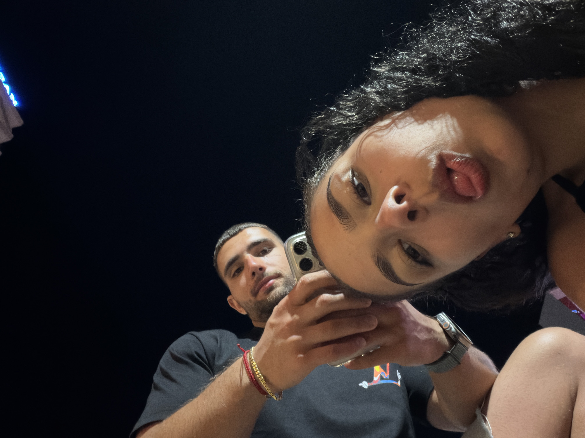 The image depicts a close-up, low-angle selfie taken at night, presumably in Nesebar, Bulgaria, given the context. Two individuals are visible. In the foreground, a woman is facing the camera with her tongue sticking out, her eyes partially closed and hair dark and curly. Behind her, a man is holding a smartphone, its camera pointed towards the subjects. He is looking down at the camera. The background is dark, suggesting an outdoor night setting. Hints of illuminated signage with blue lettering are visible at the top, possibly indicating a commercial area or attraction in Nesebar. The man is wearing a black t-shirt and several bracelets on his wrist. The woman's skin appears smooth, and she is wearing earrings.