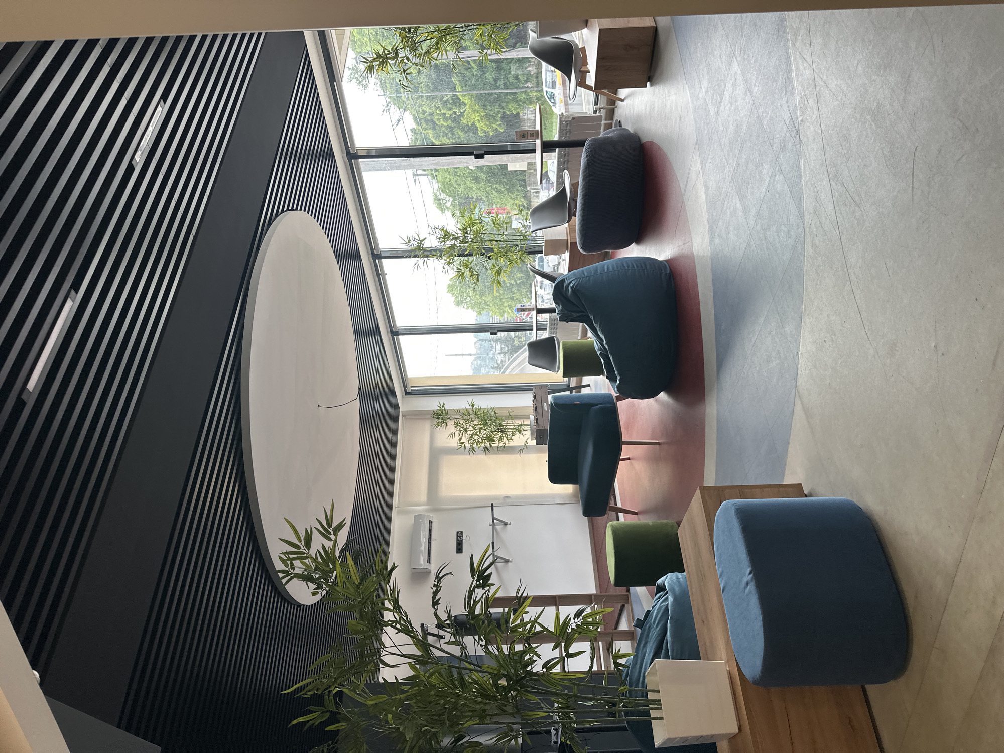 The image depicts the interior of a modern lounge area, likely a coworking space or cafe, located in Sochi, Russia. The space features a contemporary design with a black slatted ceiling and a large circular white ceiling feature. Expansive windows overlook an outdoor scene with greenery and a street, suggesting an urban environment.
Inside, the seating arrangement includes several comfortable pieces: a teal armchair with metal legs, large teal and dark grey beanbag chairs, and a blue oval ottoman. A green cylindrical ottoman is positioned near the armchair. A wooden rectangular table with a glass top is visible, accompanied by three modern grey chairs. A wooden chest or bench is placed to the left of the frame, holding a white planter with a tall green plant, possibly bamboo. Another tall green plant stands to the right, near the window.
On the back wall, an air conditioning unit is mounted, and a wooden structure with metal bars, resembling a pull-up bar, is affixed to the wall. A punching bag hangs from the structure. Further to the right, another tall green plant is visible next to a foosball table.
The floor has a unique design, with colored sections of red, grey, and off-white. The overall atmosphere is relaxed and inviting, designed for casual seating and possibly work or recreation. The natural light filtering through the windows creates a bright and airy feel.