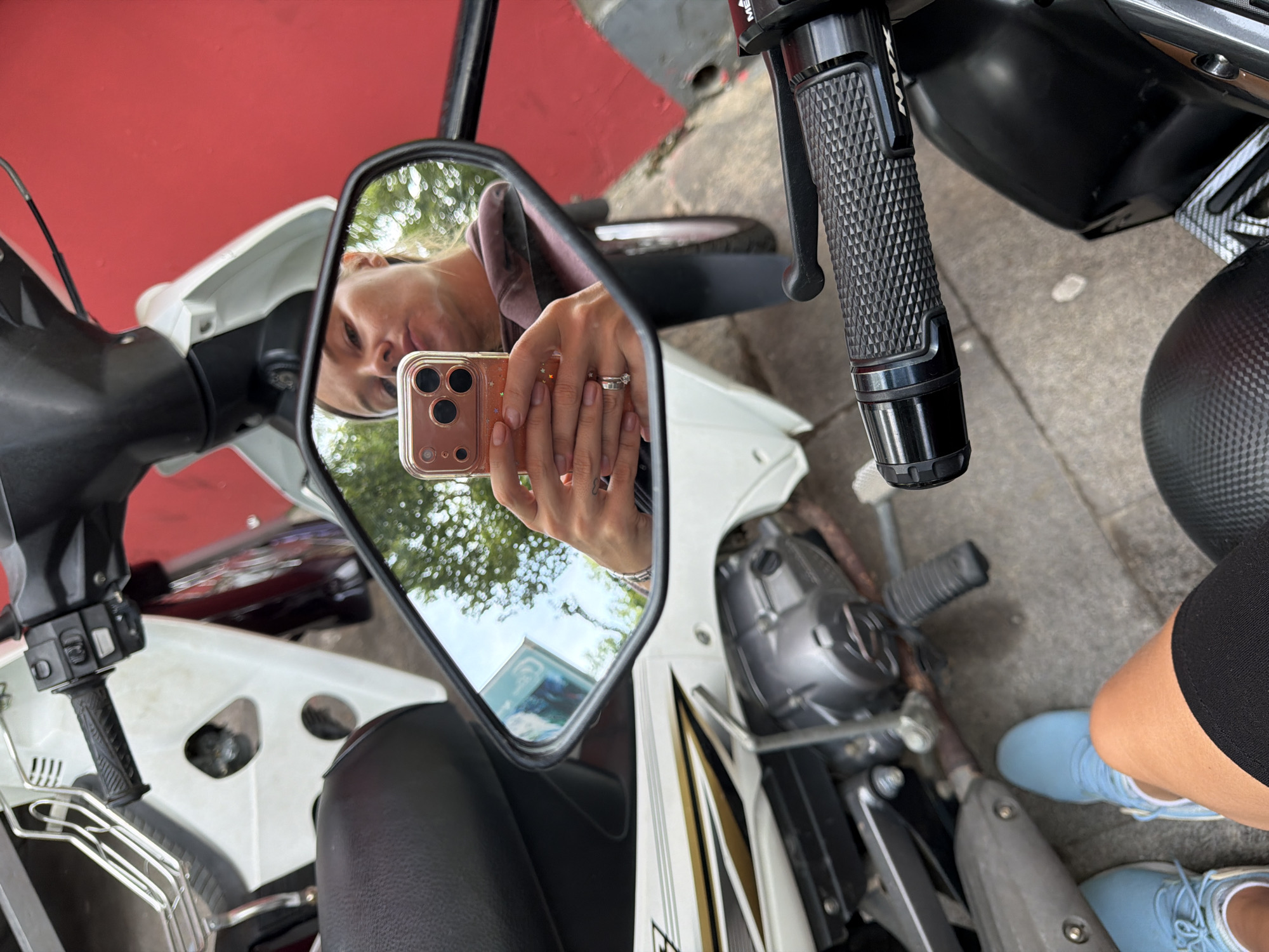 In Da Lat, Vietnam, a person takes a mirror selfie with a smartphone while sitting on a white and black motorbike. The reflection shows the person's face and hand holding the phone. The surrounding environment includes a red wall, trees, and the ground. The motorbike has a black seat, handlebars, and footpegs. The person is wearing light blue sneakers and a black skirt.