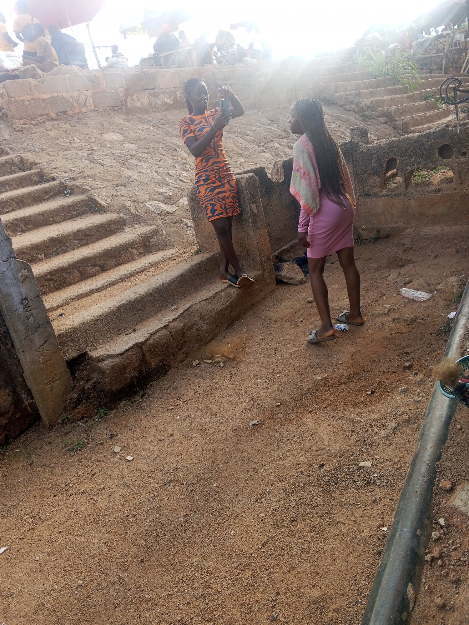 Two young women are standing outdoors in a public space. One woman is wearing an orange and black patterned dress and is holding a smartphone, appearing to take a photo. The other woman is wearing a pink dress and a light shawl or scarf. They are standing on a dirt ground near a set of concrete stairs. In the background, there is a stone structure with additional stairs leading upwards, with people visible at the top. The location appears to be in a city, possibly Gwong, Nigeria. The lighting suggests it is daytime, possibly late morning or early afternoon. The overall scene suggests a casual, informal setting, likely a public area in a city or town.