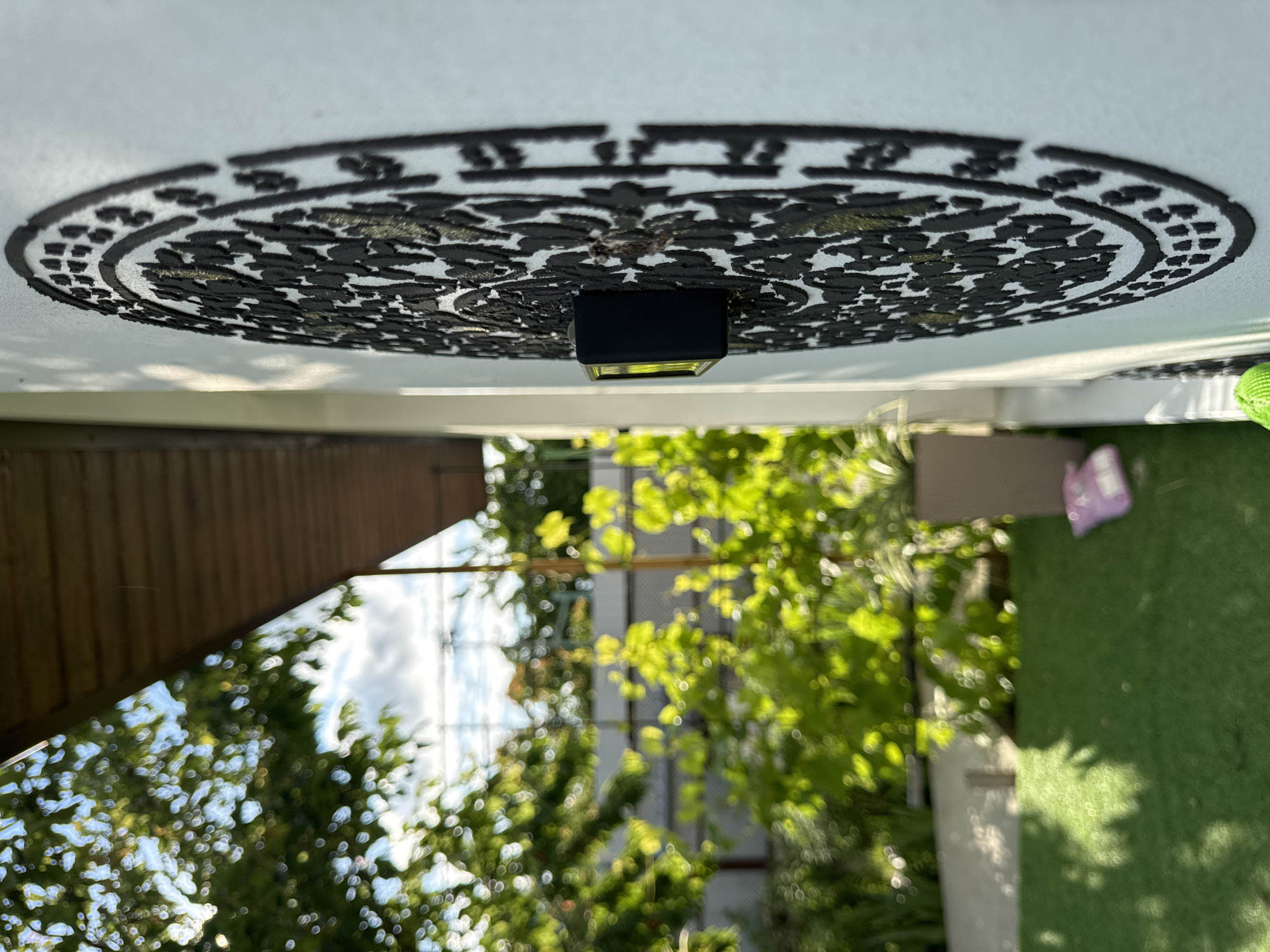 An outdoor shot, taken at a low angle, focuses on a textured, circular black decorative piece mounted on a white wall. The decoration features an intricate floral or paisley pattern with some golden accents. A small, rectangular black object with a yellow-green illuminated screen is attached to the center of the decoration. The wall appears to be part of a building with an overhanging wooden roof visible at the top left. To the left and out of focus, a garden area is visible, with lush green foliage, including trees and vines, bathed in sunlight. The ground is covered with artificial green turf, and a light pink item is visible on the turf in the middle ground. The background shows a chain-link fence and more greenery. The image location is stated to be Dobrovske rural settlement, Ukraine, suggesting a rural or suburban setting. The time of day appears to be daytime, given the bright lighting and shadows. There are no people visible in the frame.
