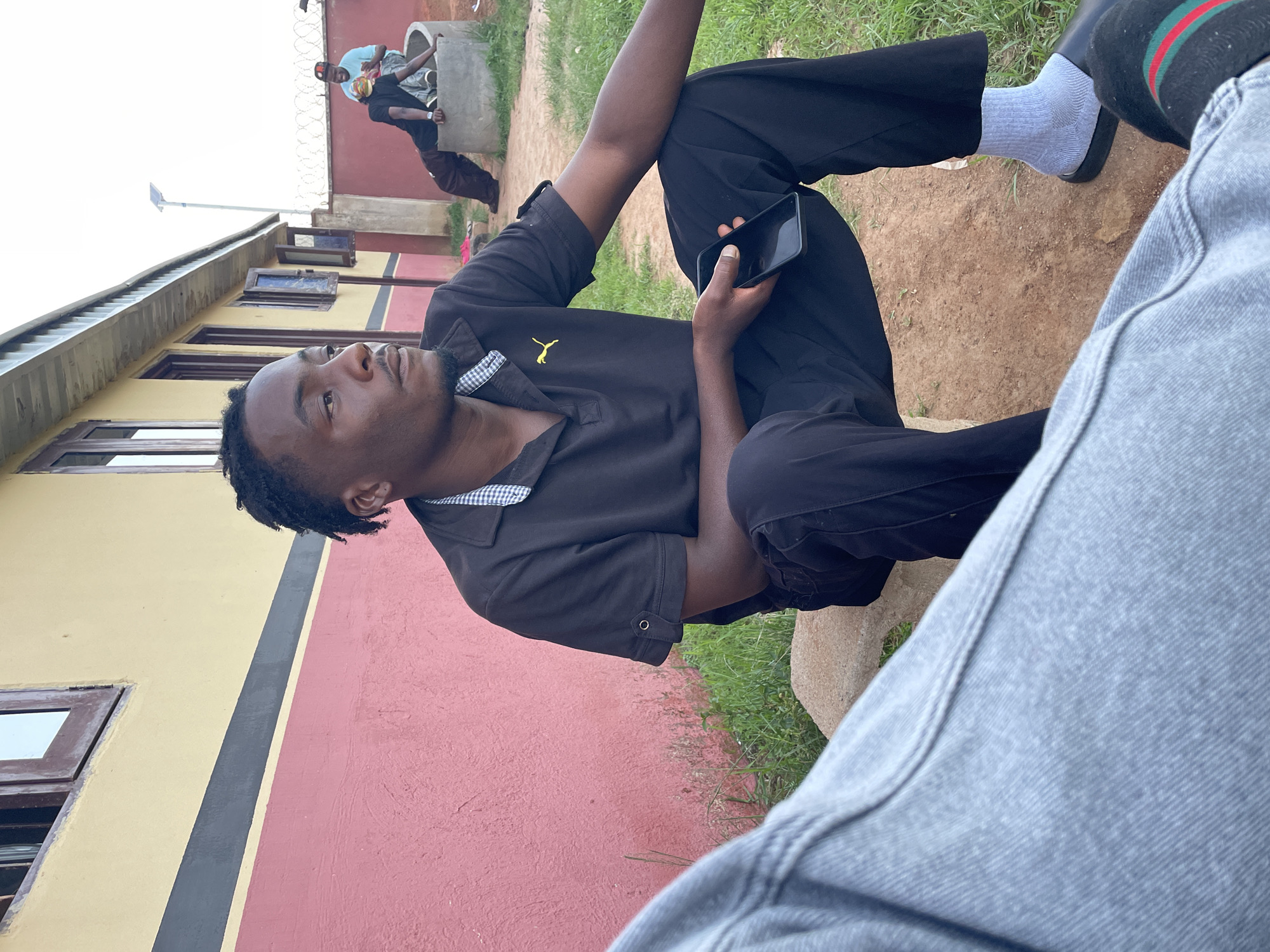 A man sits outside with a mobile phone in his hand. He wears a black polo shirt and black trousers. Another man is sitting close by. In the background, two men are near a concrete structure. The building behind the main subject is painted yellow and pink. The picture was taken in Durimi, Nigeria.