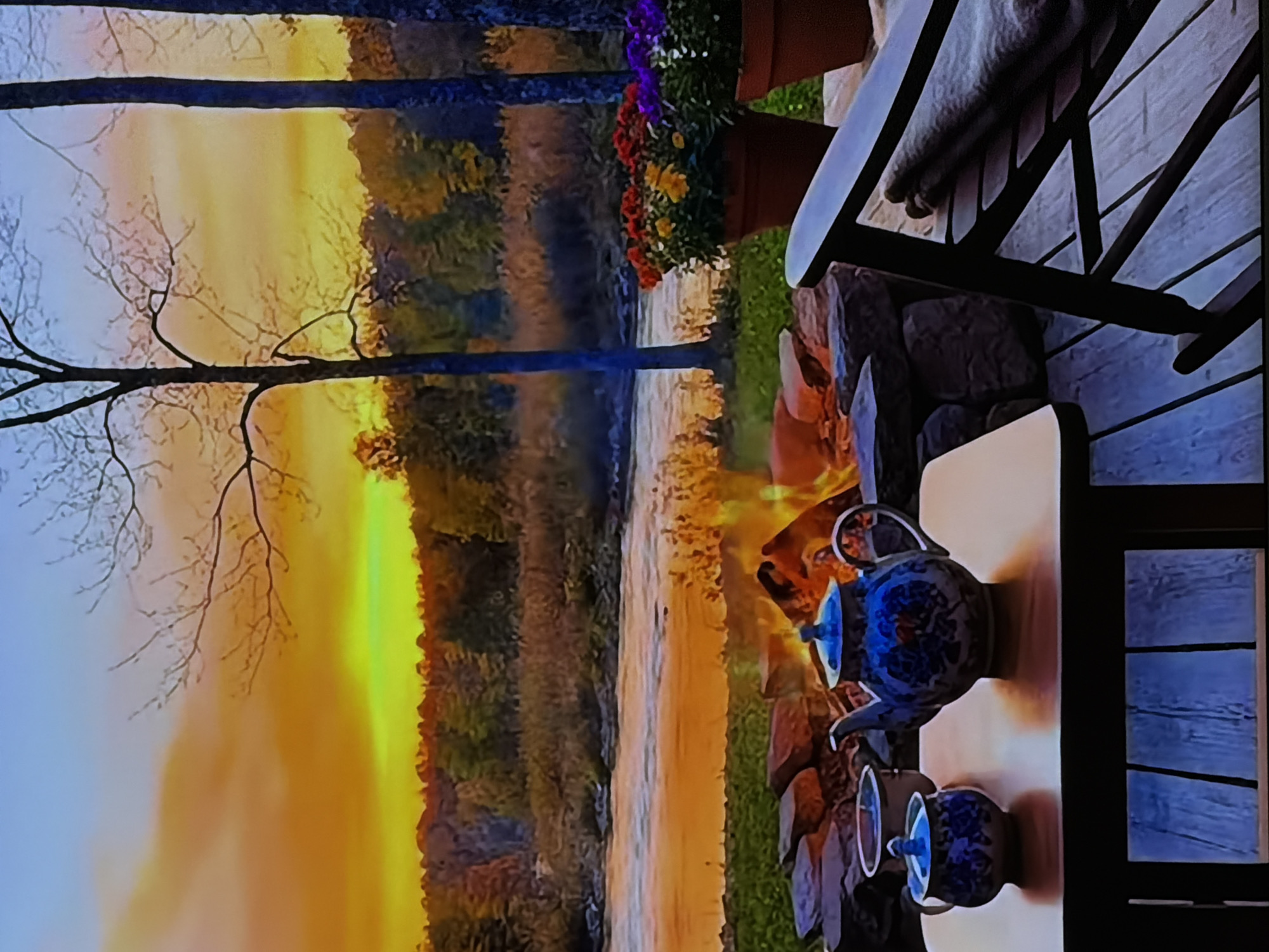 This image captures a tranquil outdoor scene at sunset, likely in Sofiivska Borshchahivka, Ukraine. The setting sun casts a warm, vibrant orange and yellow glow across the sky, which is reflected softly on a body of water, possibly a river or lake, that stretches across the middle ground. A dense treeline or forest silhouettes the far bank of the water. On the left side of the foreground, a prominent tree with delicate bare branches stands against the colorful sky.
To the right, a cozy outdoor seating area is arranged. A fire pit with visible flames provides warmth and a focal point for the gathering. Next to the fire, a low, light-colored table holds a beautiful blue and white patterned teapot accompanied by two matching cups, suggesting a tea or hot beverage serving. A wooden chair or bench, partially visible further right, has a blanket draped over it, implying comfort and relaxation. Behind the seating area, colorful potted flowers in shades of red, purple, and yellow add bursts of vibrancy to the scene.
The overall impression is one of peace, relaxation, and warmth, capturing a serene evening spent by the fire enjoying the natural beauty of the sunset. There is no visible text in the image.