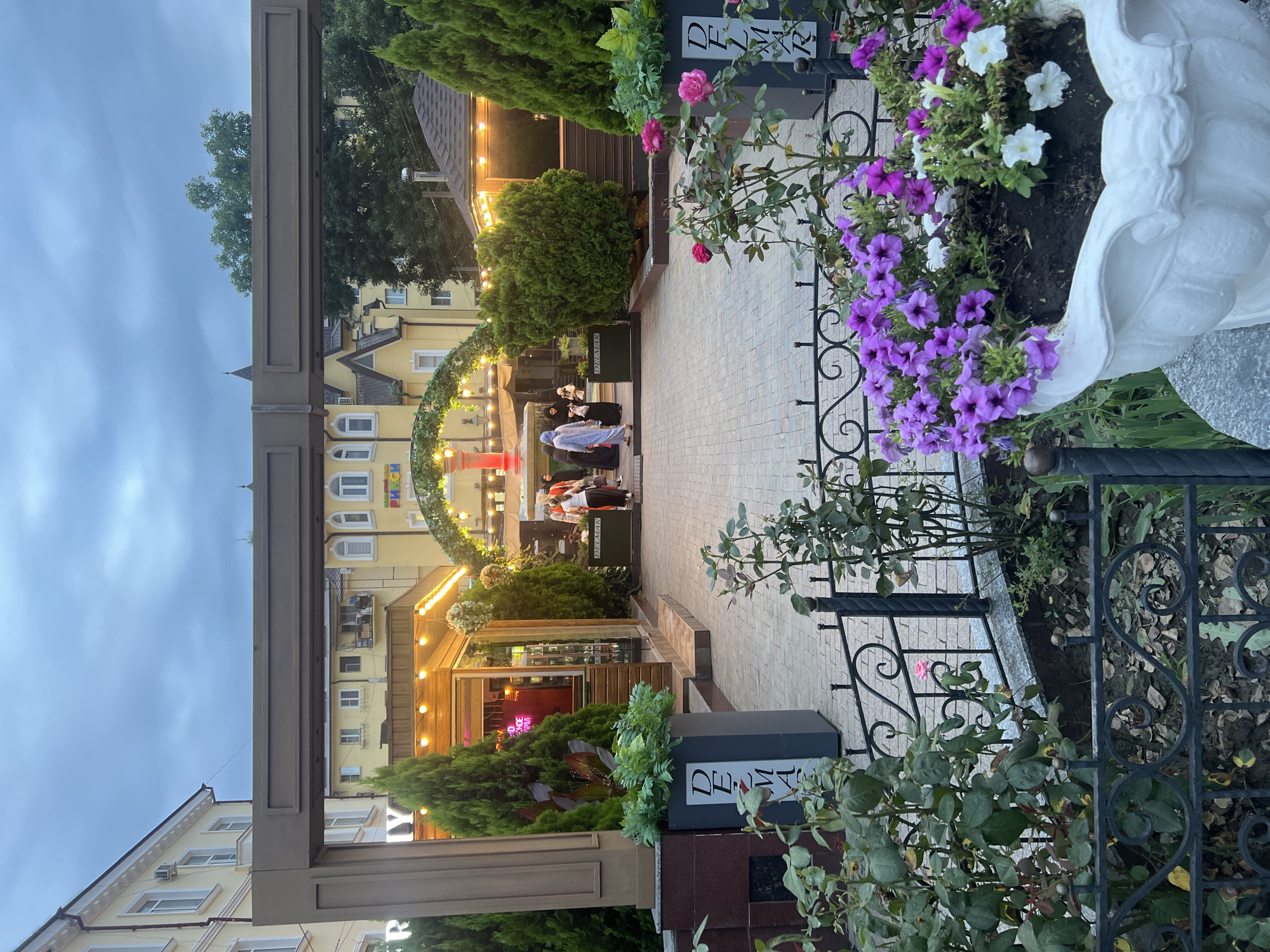 The image depicts an outdoor dining area or courtyard in Grozny, Russia. A group of people, some in traditional Islamic attire, are seen walking towards an arched entrance decorated with greenery and lights. The architecture of the surrounding buildings is light yellow with white trim and arched windows. There are several potted plants, including cypress trees and flowering bushes, as well as decorative flower pots filled with petunias and roses in the foreground. A dark brown archway frames the entrance to the courtyard. The sky is overcast, suggesting it is either early morning or late evening.