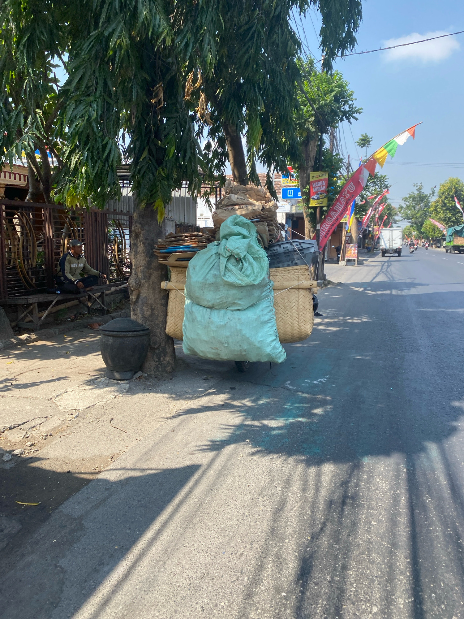 A medium shot shows a street scene in Kediri City, Indonesia, on a sunny day. The foreground is dominated by a two-wheeled cart loaded with a large, light green tarp-covered bundle and several woven baskets filled with various items. Behind the cart, a person sits on a wooden bench next to a large black pot. The person is wearing a cap, a jacket, and pants. A large tree with drooping branches casts shadows on the ground. To the right of the cart, the street extends into the distance, with several shops and buildings lining the road. Indonesian flags and banners are visible along the street, suggesting a possible celebration or event. A white truck and several motorcycles are on the road, indicating moderate traffic. The environment appears to be an urban or semi-urban area, with a mix of natural elements like trees and man-made structures.