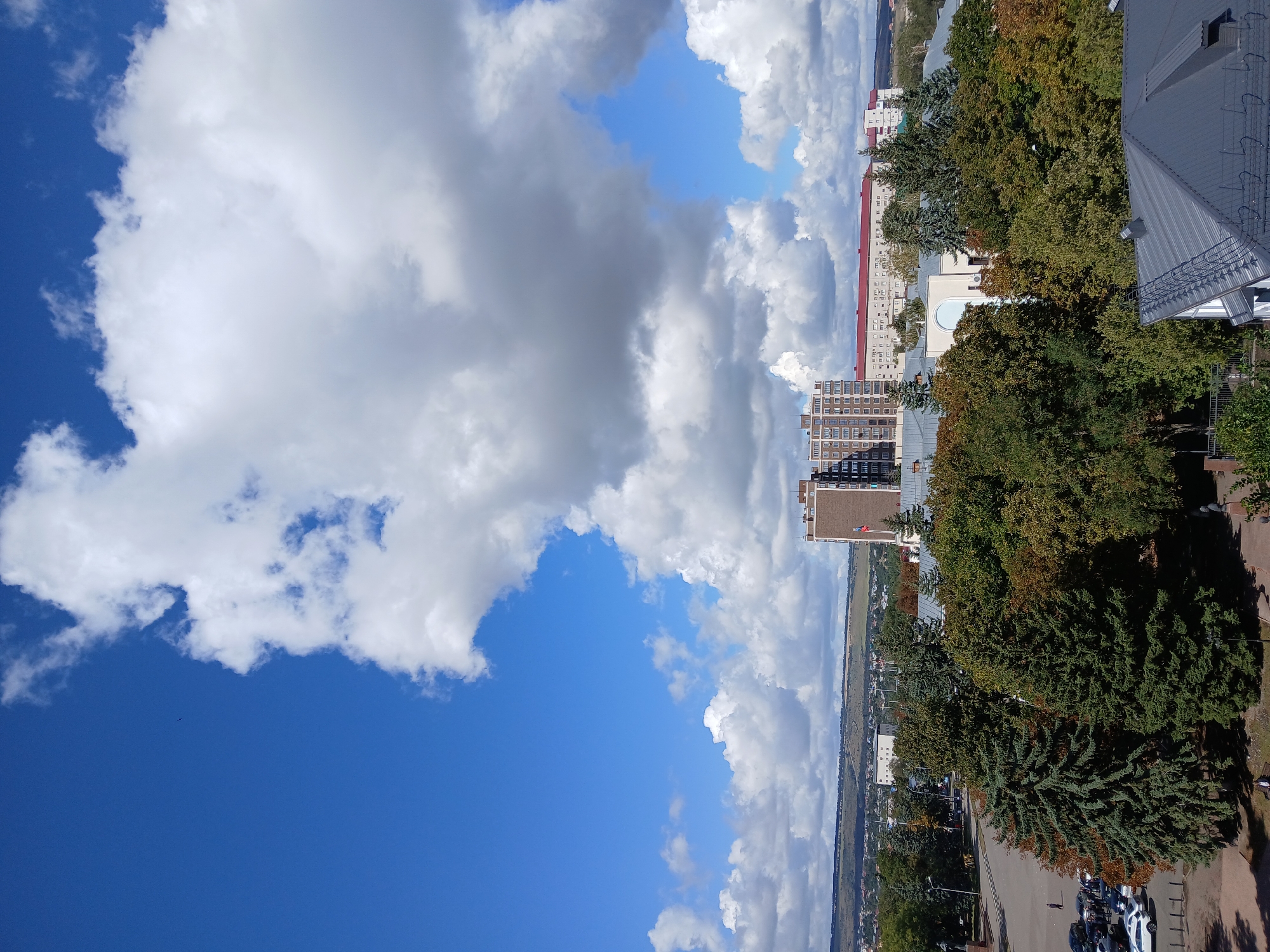 The image depicts a panoramic view of a city with buildings and trees under a cloudy sky. In the foreground, several cars are parked on a paved area, with a few people visible. A tall, modern building with a brick facade stands prominently in the middle ground, along with a longer, lower building with a red roof. Lush green trees, some with autumn foliage, fill the space between the buildings and the foreground. In the background, a distant landscape with rolling hills is visible. The sky is a vibrant blue with large, fluffy white clouds. This scene is likely depicting a central area of Cherkessk, Russia, potentially near a public square or park given the open space and presence of multiple buildings and trees.