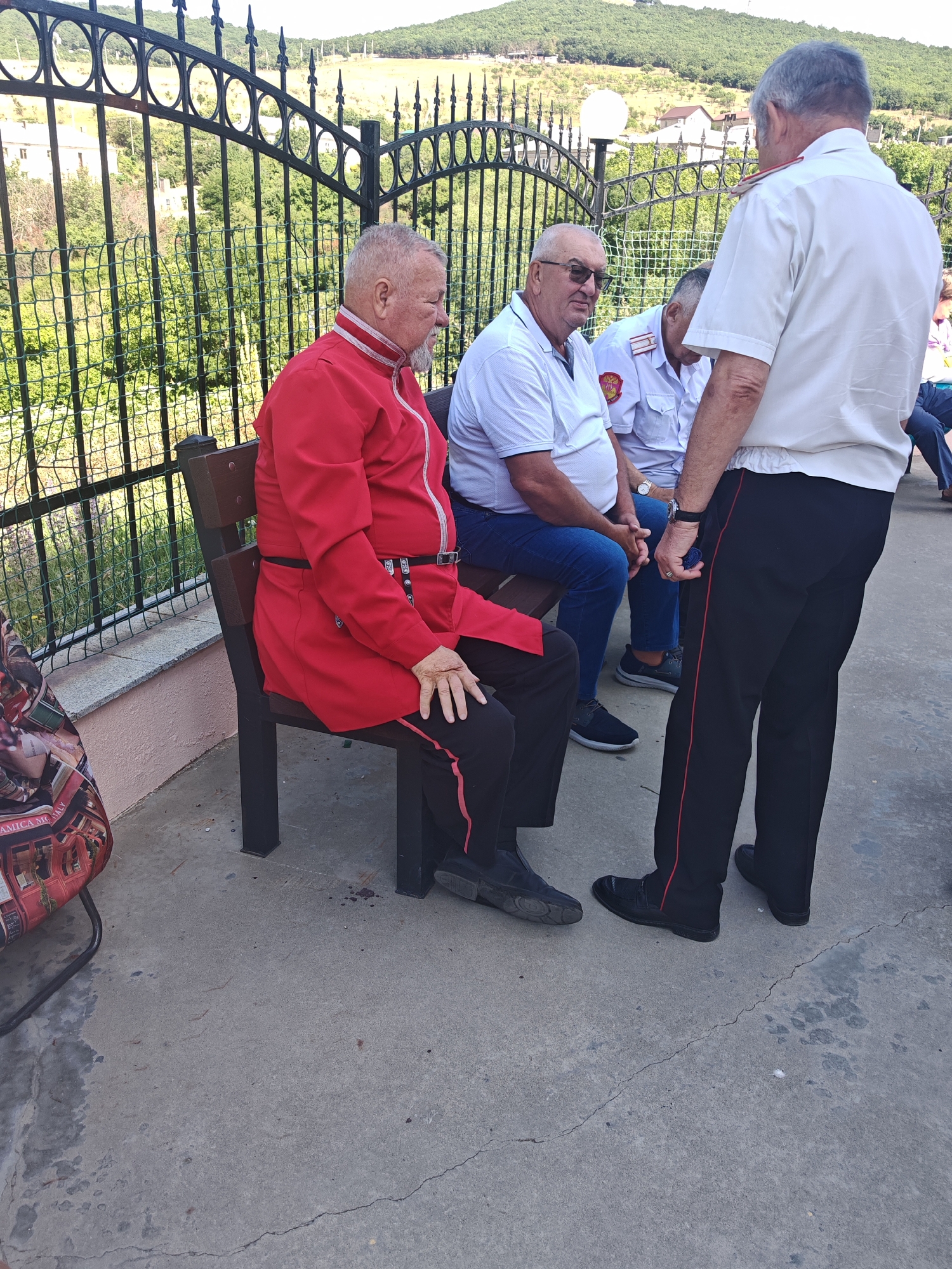 In Varvarovka, Russia, three men are gathered outdoors. One man, seated on a bench, wears a red uniform jacket with ornate trim, black pants, and a black belt. He has a grey beard and is looking to his right. Another man, also seated, is wearing a white polo shirt and blue jeans, with glasses and a watch. He is looking towards the first man. The third man stands, facing the seated men, dressed in a white shirt with red trim on the collar and black pants with a red stripe. He is holding a small, round, blue object. Behind them is a black metal fence, and beyond that, a hillside covered in trees and some buildings. The ground is concrete, with some scattered debris. A patterned bag rests on the ground to the left of the bench.