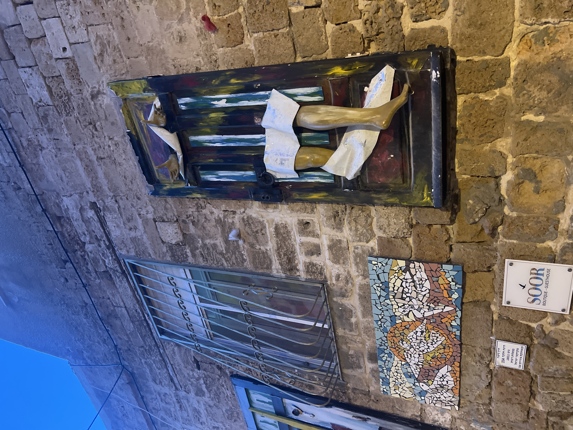 The image captures a vibrant section of an old stone wall, likely in a narrow alleyway or street in Acre, Israel, given the architectural style and visible text. The wall is constructed from large, irregular, weathered stone blocks, showcasing historical craftsmanship. The scene is illuminated by daylight, with a clear blue sky visible towards the bottom left, suggesting good weather.
Several artistic and functional elements adorn the wall. At the top, a unique art installation resembles a distorted, brightly painted wooden frame or door, from which golden-colored human-like limbs—a pair of hands and a pair of legs/feet—protrude, partially wrapped in white fabric. This creates an intriguing, somewhat surreal, visual effect.
Below this installation and to its left, a window with an ornate metal security grill is set into the stone wall. The grill features decorative scrollwork.
Further down and to the right, another piece of art is displayed: a rectangular mosaic, intricately crafted from small tiles in shades of blue, orange, brown, and white. The mosaic depicts an abstract design, possibly stylized birds or doves.
Two signs are mounted next to the mosaic. The upper sign, in English, reads "SOOR / BOUTIQUE - GUESTHOUSE" and features a small bird silhouette, indicating a local business. Below it, a white rectangular sign with a downward-pointing arrow contains Hebrew text, which translates to: "Access road / is parcel 334 / 18011 in block / owned by the Israel Land Authority / and maintained by the Acre Municipality," providing specific details about the property. Thin black electrical wires are also visible running along the wall. The overall scene suggests a place rich in history, culture, and artistic expression.