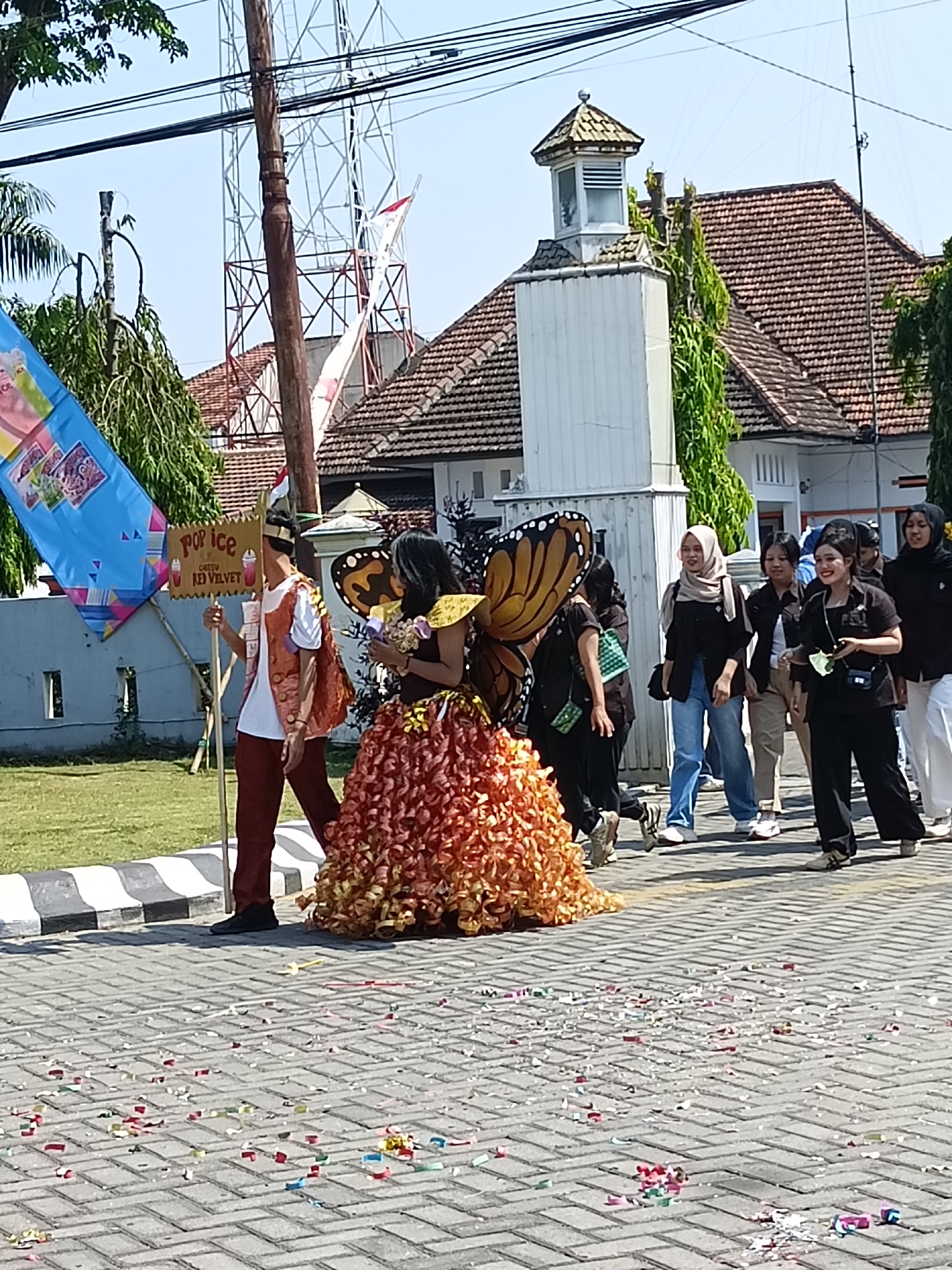 A group of people are walking on a paved street in Kediri City, Indonesia. The person in the foreground is dressed in a costume resembling a butterfly with large orange wings. They are wearing a fluffy orange and yellow skirt. To the left of the butterfly-costumed person is another person holding a flag with the words "POP ICE" and "RED VELVET" written on it. The people are walking from left to right. In the background, there is a building with a tiled roof and a tower. The street is covered in confetti.