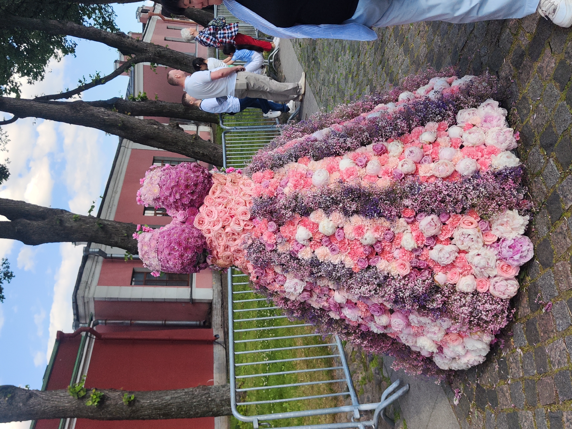 The image depicts a large, ornate floral sculpture resembling a ballgown, displayed outdoors on a cobblestone path. The sculpture is constructed from numerous pink and purple flowers, primarily roses, peonies, and smaller clustered blossoms. The dress-like form has a bodice and a voluminous skirt that fans out. In the background, a red building with white trim and arched windows is visible, suggesting an urban setting with historical architecture. Several trees with bare branches stand behind the sculpture and the building. To the right of the floral artwork, a few individuals are observed standing and walking, including a man in a white t-shirt and light-colored pants, a woman in a plaid shirt, and another person wearing a blue striped shirt and light blue pants. A metal fence runs along the left side of the image, separating the display area from a grassy patch with scattered white flowers. The sky is partly cloudy with blue patches. The overall impression is an art installation in a public space within Saint Petersburg, Russia, possibly during a festival or exhibition given the elaborate floral display.