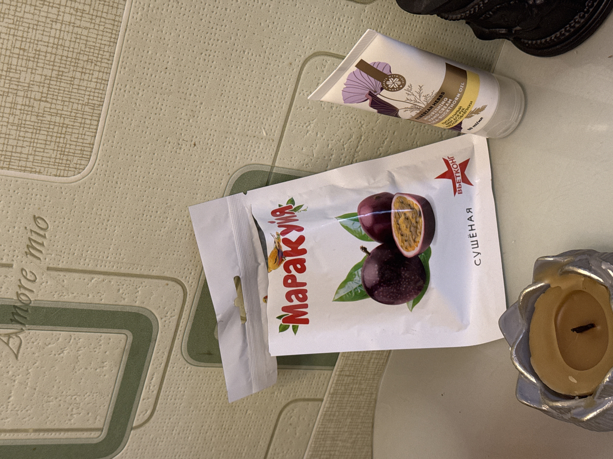 A flat-lay image shows several items on a surface. The top right corner contains a black, ornate object. Below it and angled towards the upper right is a white tube with beige and purple floral designs, labeled with the text "SIBERIAN HERBS NOURISHING HAND CREAM SEA-BUCKTHORN OIL". To the left and slightly lower is a white package with a picture of passion fruit and the word "Маракуйя" in red Cyrillic script. Beside this packet to the right sits a silver candle holder with a lit candle. The surface has a light green patterned design, and "Amore mio" is partially visible in the bottom left. There is no location specified in the city of Ketoshi, Russia, nor any people or interactions visible in the image.