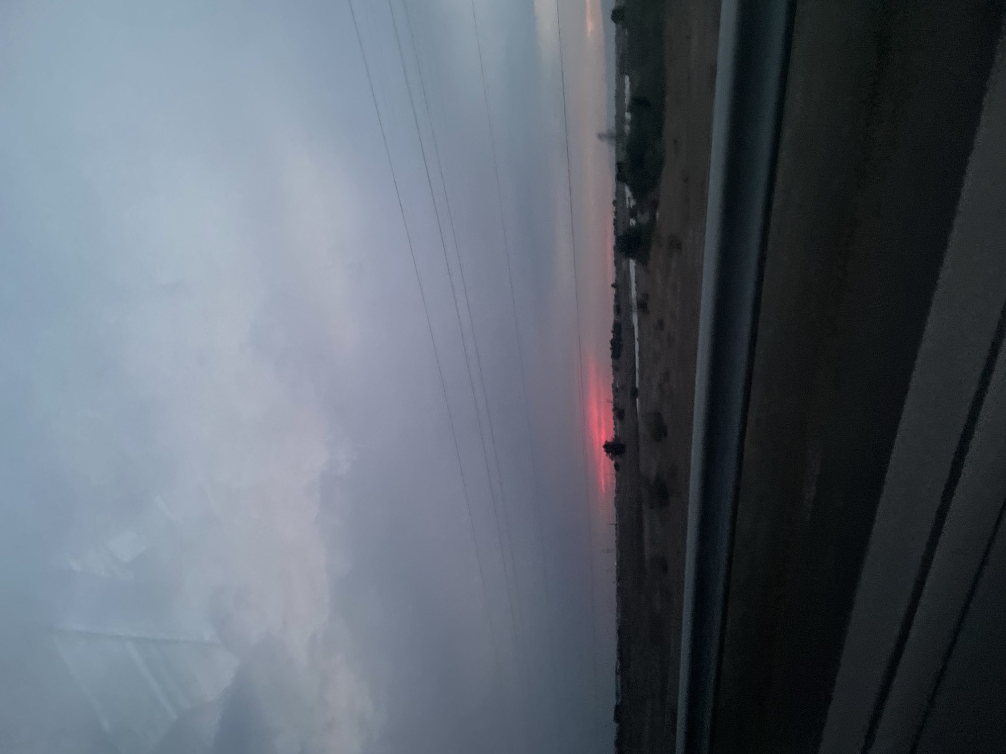 The image shows a wide-angle view of a rural landscape at dusk. A strip of dark, reflective material, possibly a road or a body of water, runs across the bottom of the frame. Above this, a sparse, dry field stretches to the horizon, dotted with low bushes and a few silhouetted trees. In the distance, a vibrant band of pink and red light indicates a sunset or sunrise, breaking through a heavy, grey, overcast sky. Several thin, dark power lines are strung horizontally across the upper portion of the sky. The overall impression is one of a vast, somewhat desolate, open area under a dramatic sky. The location is identified as Қазтай Ұлтарақов ауылы, Kazakhstan.