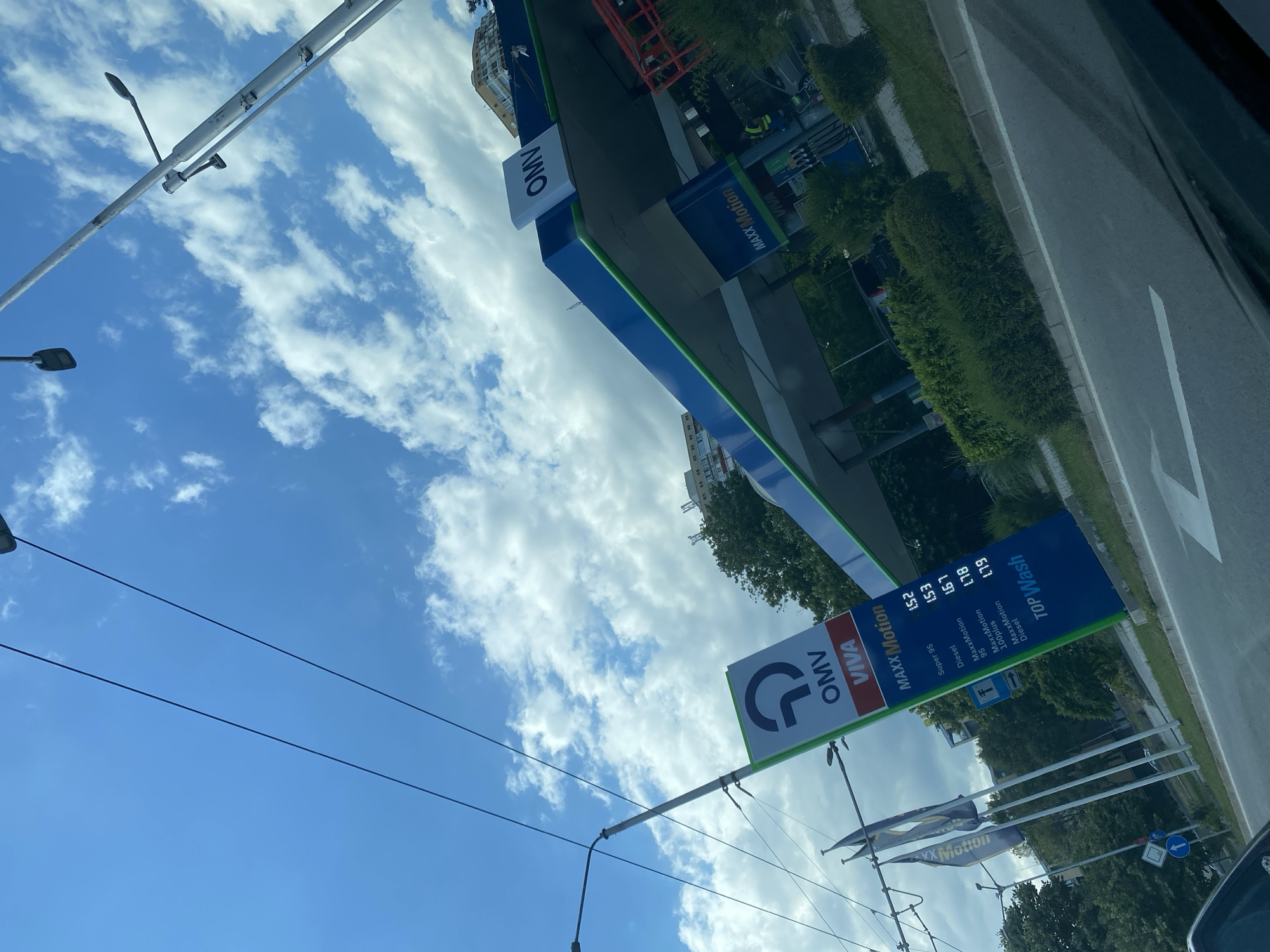 The image captures a sunny day at an OMV gas station in Varna, Bulgaria. The sky is a brilliant blue, dotted with fluffy white clouds. The perspective is from inside a vehicle, looking out at the gas station.
Prominently featured is a blue and green OMV sign with the "VIVA" logo and "MAXX Motion" branding. Below this, a price board displays fuel prices: "Super 95" at 1.52, "Diesel" at 1.53, "MaxxMotion 95" at 1.67, "MaxxMotion" at 1.78, "100plus" at 1.79, and "MaxxMotion Diesel" also at 1.79. The "TOP Wash" service is also advertised.
Another OMV sign with the circular logo is visible further to the left. Streetlights with distinct modern designs are also present. The scene includes green, well-maintained bushes lining the pavement and the road. In the background, buildings are visible, suggesting an urban or semi-urban setting. The overall impression is of a typical daytime scene at a well-kept service station, under clear, pleasant weather.