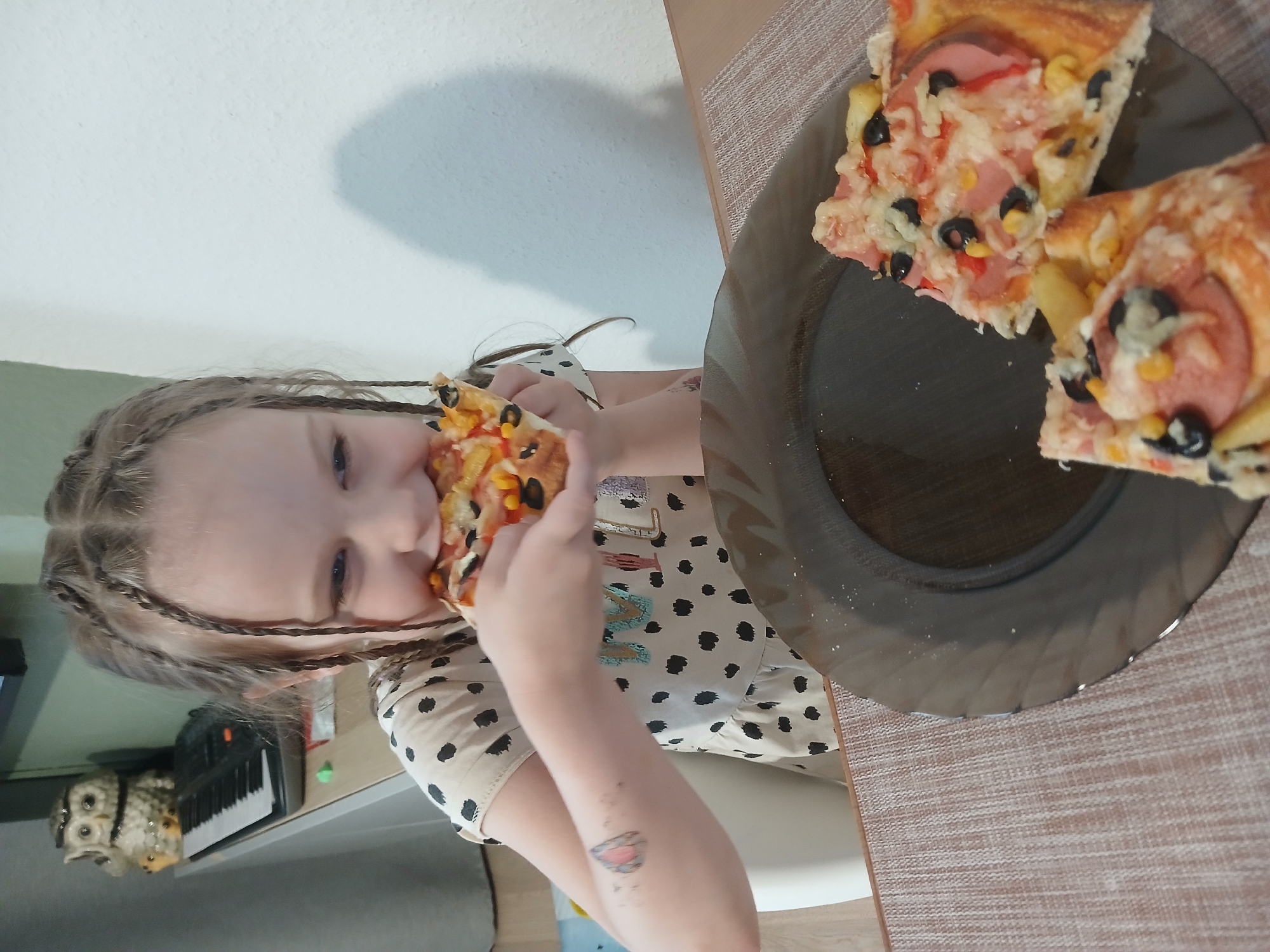 In Yekaterinburg, Russia, a young girl with braided hair is eating a slice of pizza. She is wearing a beige dress with black polka dots. A plate with two more slices of pizza sits on a table in front of her. In the background, a keyboard and an owl figurine are visible. The girl appears to be enjoying her meal.