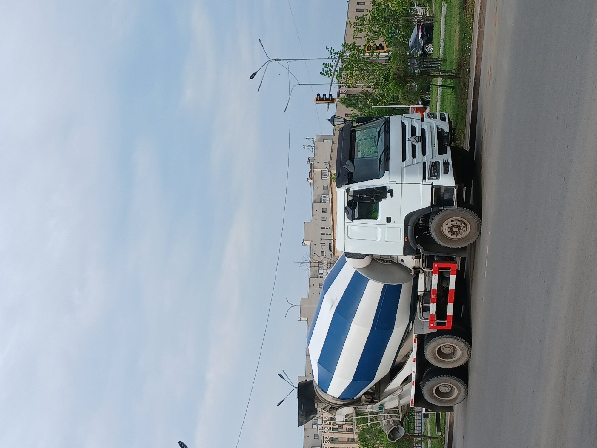 A white concrete mixer truck with blue and white striped drum is parked on the right side of a paved road in the city of Astana, Kazakhstan. The truck is facing towards the right. In the background, there are multi-story buildings, trees, and a cloudy sky. Traffic lights and street lamps are also visible. The scene appears to be an urban setting. There are no people visible in the image.