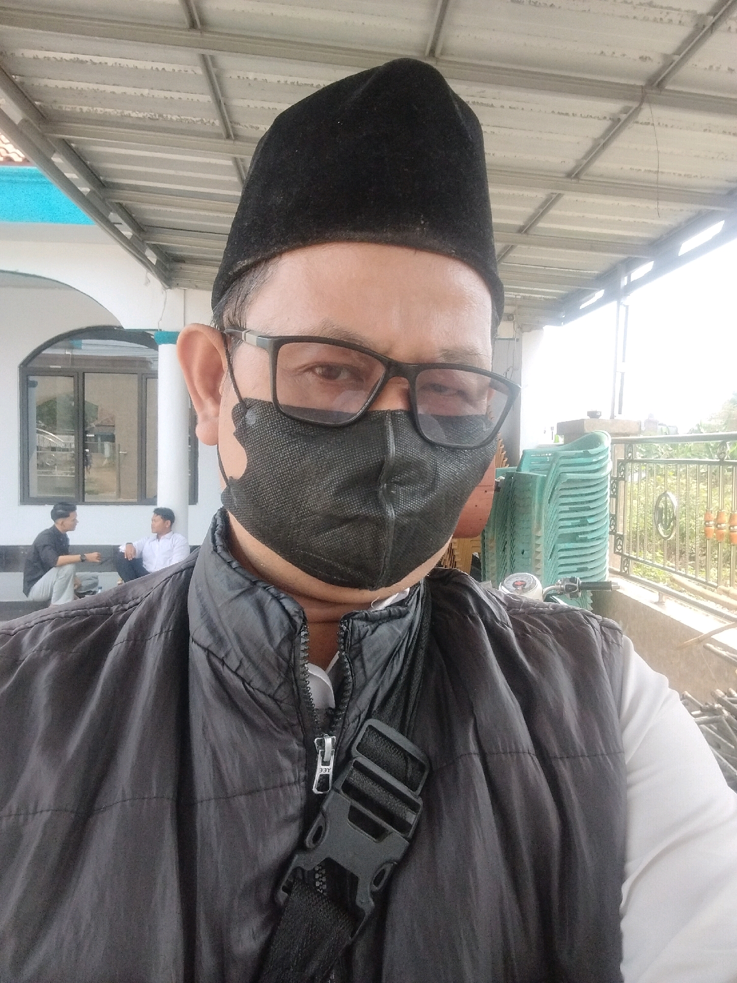 A man in his 40s, wearing a black peci (a traditional Indonesian cap), glasses, and a black face mask, takes a selfie outdoors. He is wearing a dark grey vest over a white long-sleeved shirt, with a black strap crossing his chest. Behind him, to the left, is a building with a white facade and a large window. Two men, one in a black shirt and the other in a white shirt, are sitting on steps in front of the building, engaged in conversation. To the right, a stack of light green chairs is visible, along with a metal railing and some foliage. The scene appears to be in Serang, Indonesia, possibly near a mosque or public gathering area, indicated by the building's architecture and the presence of the peci. The overall lighting suggests it is daytime.