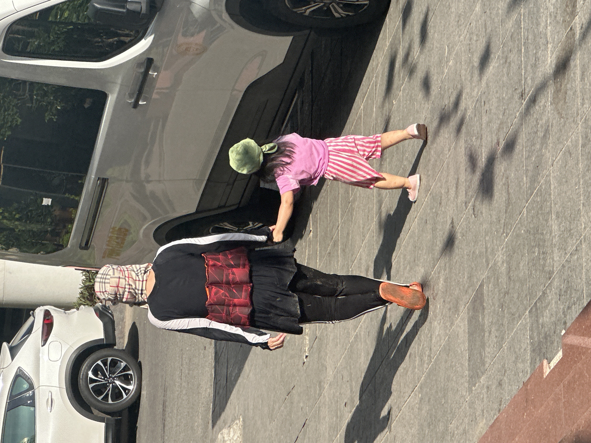 In Bac Nha Trang Ward, Vietnam, two figures walk across a paved city sidewalk under bright sunlight. A taller person, seen from behind, wears a checkered headscarf, a black long-sleeved top with white stripes on the sleeves, and a ruffled red garment over a black skirt. They also wear black leggings and orange slip-on shoes. Next to them, a small child in a pink t-shirt and pink and white striped shorts, topped with a green hat, holds the taller person's hand. Both are casting distinct shadows on the gray, square-tiled pavement. A white SUV is parked to the left, and a silver van is partially visible to the right, suggesting an urban street or parking area. The scene is captured from a slightly elevated perspective, looking down on the figures and the surrounding environment.