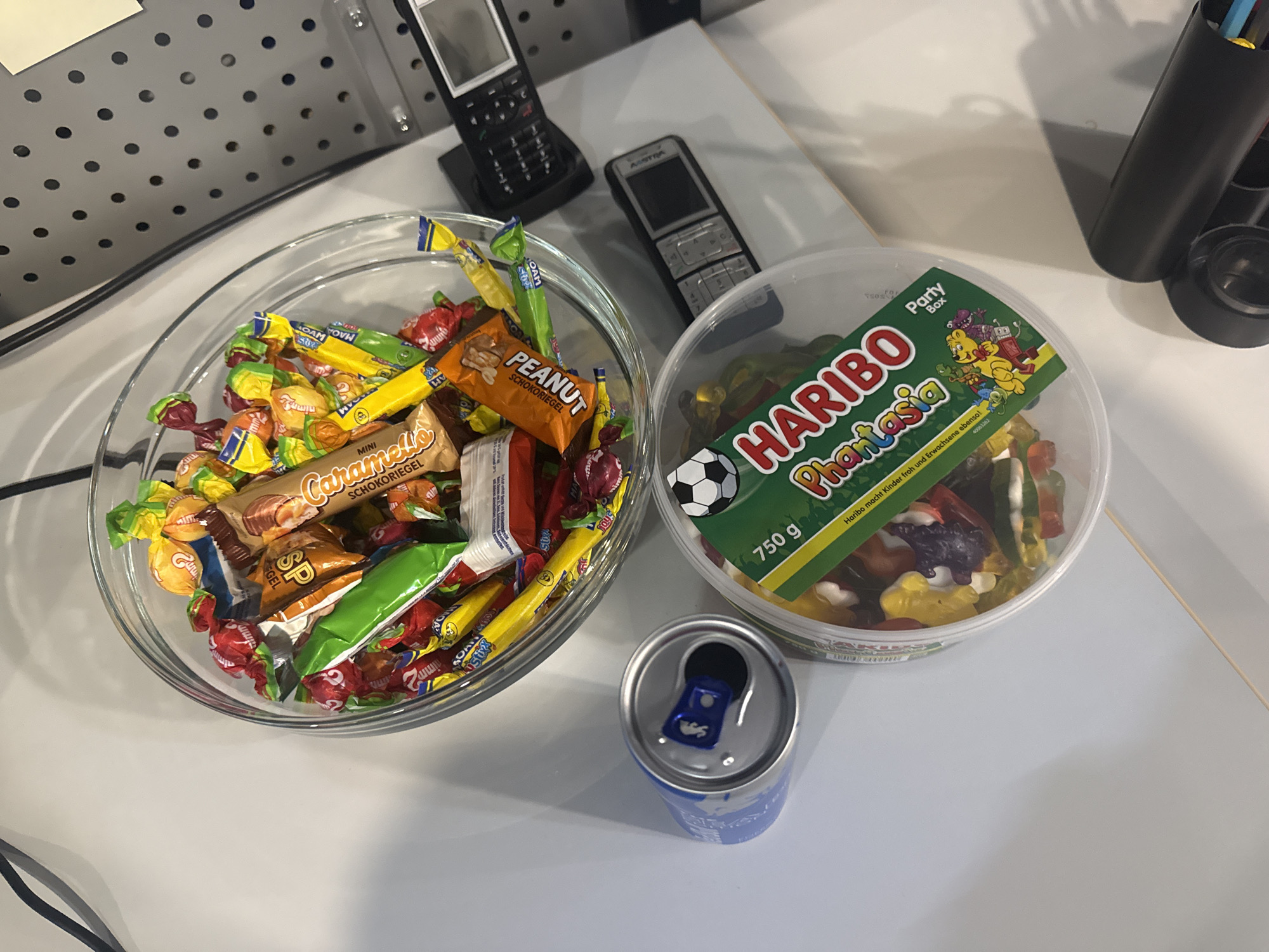 The image shows a desk with various items, including a glass bowl filled with an assortment of wrapped candies, a can of Red Bull, a clear plastic container of Haribo Phantasia gummy candies, and two mobile phones. The candies in the bowl appear to be a mix of chocolate bars and other individually wrapped sweets. One phone, a black Nokia model, is positioned on the right side of the image, and another, silver and black, is below it. The Haribo container is open, revealing a colorful collection of gummy candies. A blue and silver Red Bull can is next to the bowl. The desk surface is white, and a grey perforated board is visible in the background. The scene is viewed from above, with a shallow depth of field. The location is identified as Rehestädt, Germany.