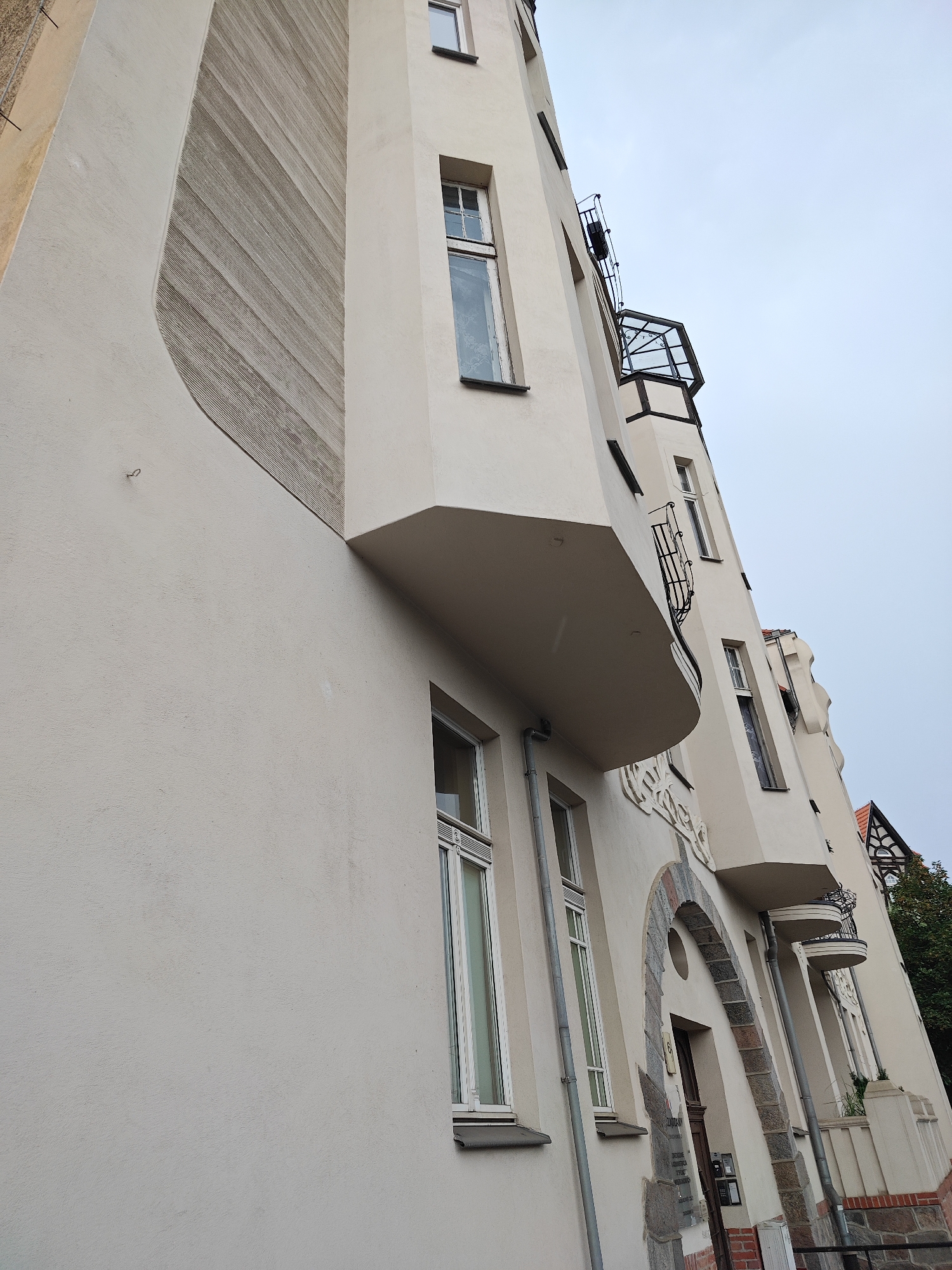 The image captures a low-angle view of a light-colored building facade with an overcast sky. The building features several windows, balconies, and a textured section on the left side. The architectural style appears to be somewhat eclectic, with a mix of modern and traditional elements. The lower portion of the building shows an arched doorway with stonework. The facade is primarily a pale, neutral color, possibly cream or beige, with some darker accents around the windows and balconies. The textured section on the left is a darker gray and has a ribbed pattern. The overall impression is of an urban environment, possibly a residential or commercial building. The specific location in Poznan, Poland, is not evident from the image itself, but the building's style might suggest a historical district.