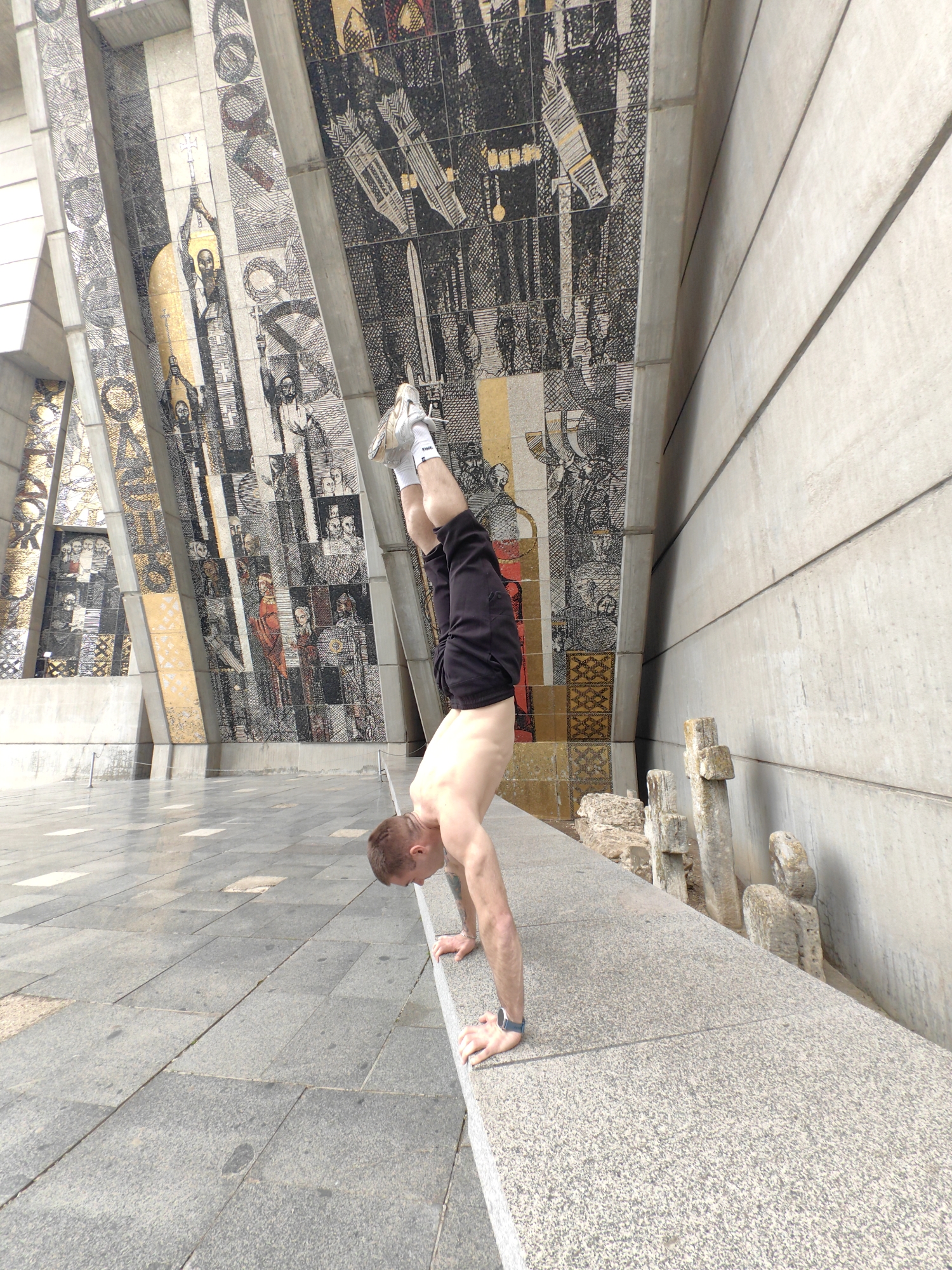 A man is performing a handstand on a ledge in front of the Founders of the Bulgarian State Monument in Shumen, Bulgaria. The man is wearing black shorts, white socks, and sneakers. He is shirtless and has a wrist watch on his left wrist. The monument consists of large mosaic figures and concrete structures. Stone crosses and rubble lie behind the ledge where the man is performing his handstand. The ground is paved with light grey stones.