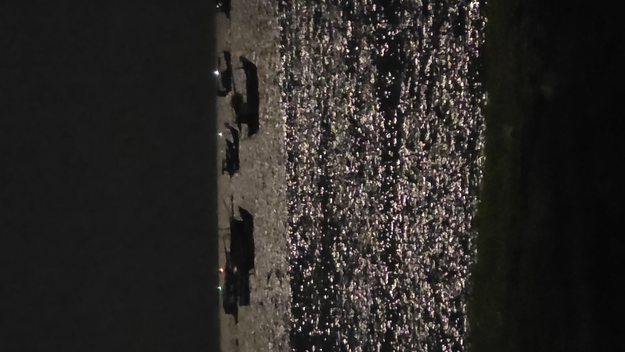 At night, the image shows a line of parked vehicles, possibly cars, illuminated by streetlights on the left. To the right of the vehicles is a body of water reflecting light. The location is likely near the coast in Bắc Nha Trang, Vietnam, given the coastal environment and presence of water. The scene appears dark with limited visibility.