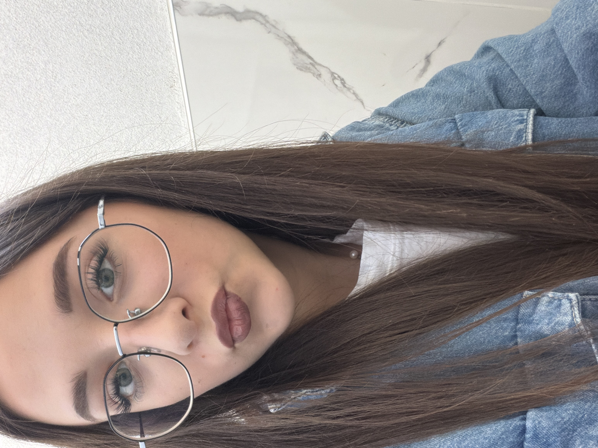 A close-up, angled, portrait-style photograph shows a young woman with long, dark brown hair wearing round, silver-framed glasses. She is wearing a white shirt or top visible at her neckline and a blue denim jacket. Her makeup includes defined eyebrows, long eyelashes, and mauve lipstick. The background consists of two different white surfaces, one possibly a tiled wall or floor with gray marbling, and the other a solid white surface. There is no evidence indicating the location is Zmeitsa, Bulgaria, or any other specific location.