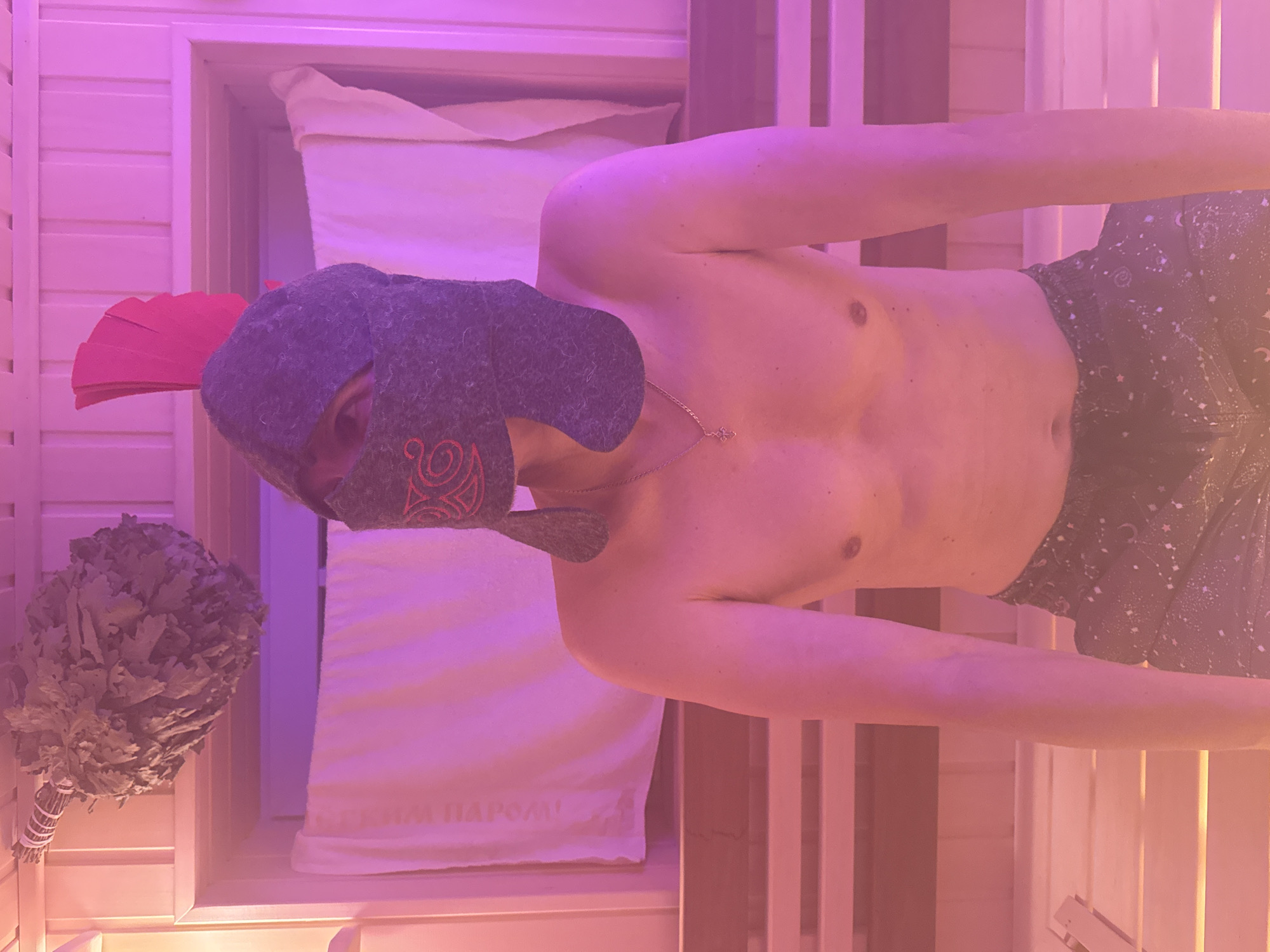 A shirtless person wearing a grey helmet with red detailing and a red plume, resembling a Roman helmet, sits on a wooden bench in a sauna. The person has a necklace with a cross pendant and is wearing patterned shorts. To the upper left, a bundle of dried branches, possibly a venik for a sauna, hangs from the ceiling. The sauna is lit with pink and purple lighting. The location is identified as Zelenaya Roshcha, Russia.