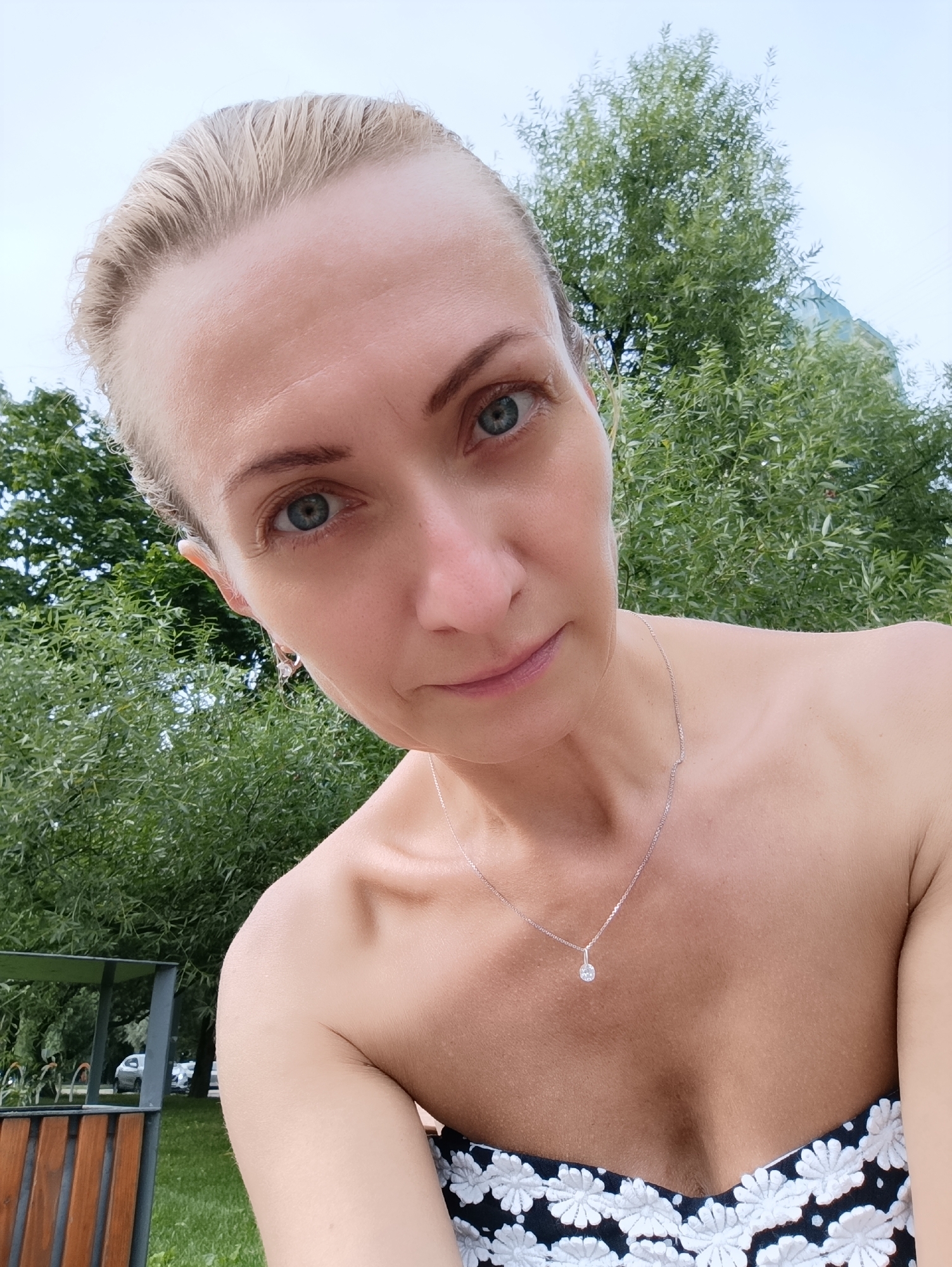 A woman with blonde hair pulled back and blue eyes looks directly at the camera in a selfie. She is wearing a strapless top with a black and white floral pattern and a delicate silver necklace with a small pendant. The background is lush green foliage, suggesting an outdoor setting. The image appears to have been taken in Moscow, Russia, based on the provided context.