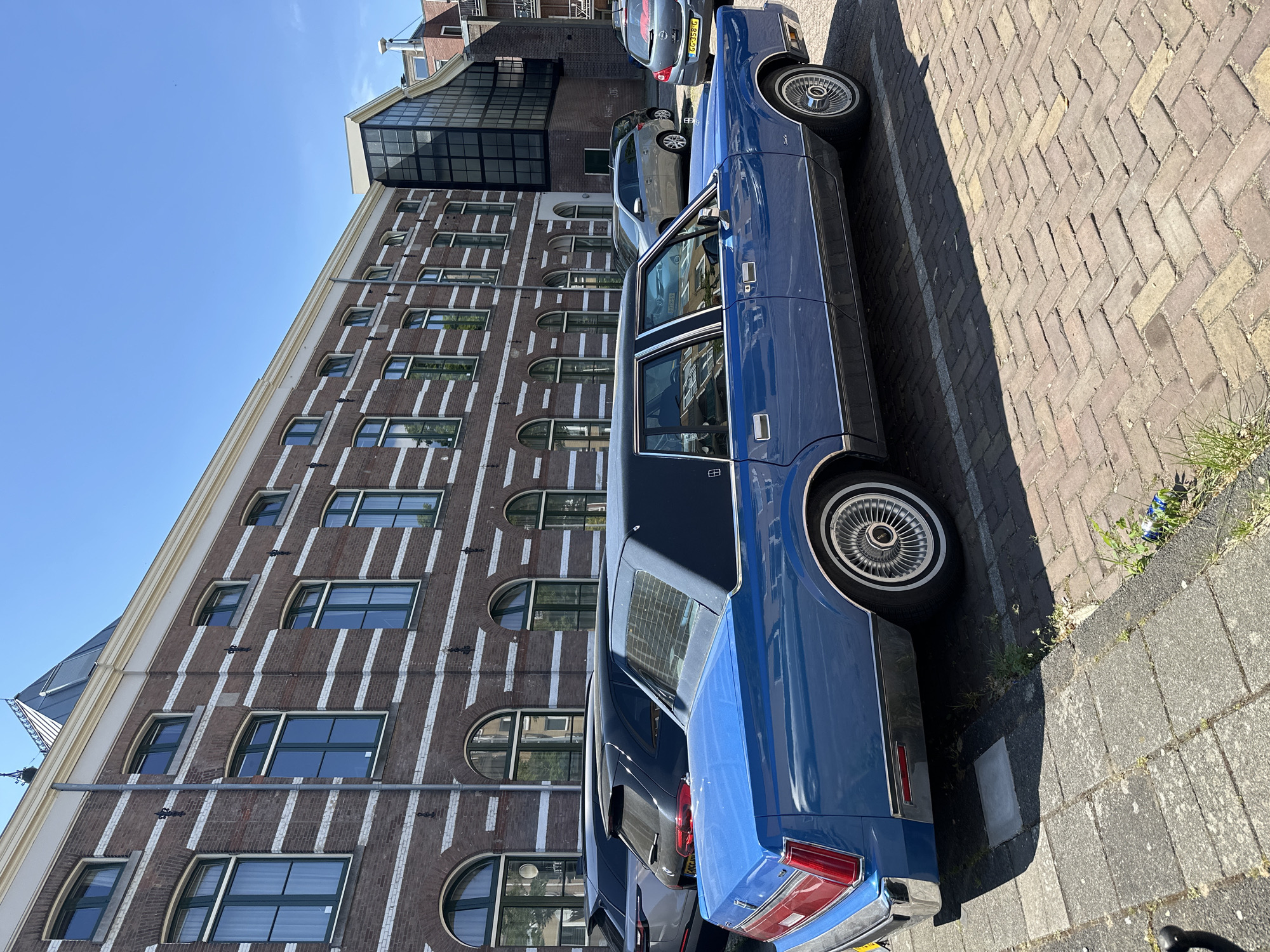 A blue Lincoln Continental Town Car is parked along a brick sidewalk in Amsterdam, Netherlands. Adjacent to the car is a row of parked vehicles. The background features a building with a distinctive brick façade, accentuated by vertical white lines and arched windows. The sky is clear and blue, suggesting a sunny day.