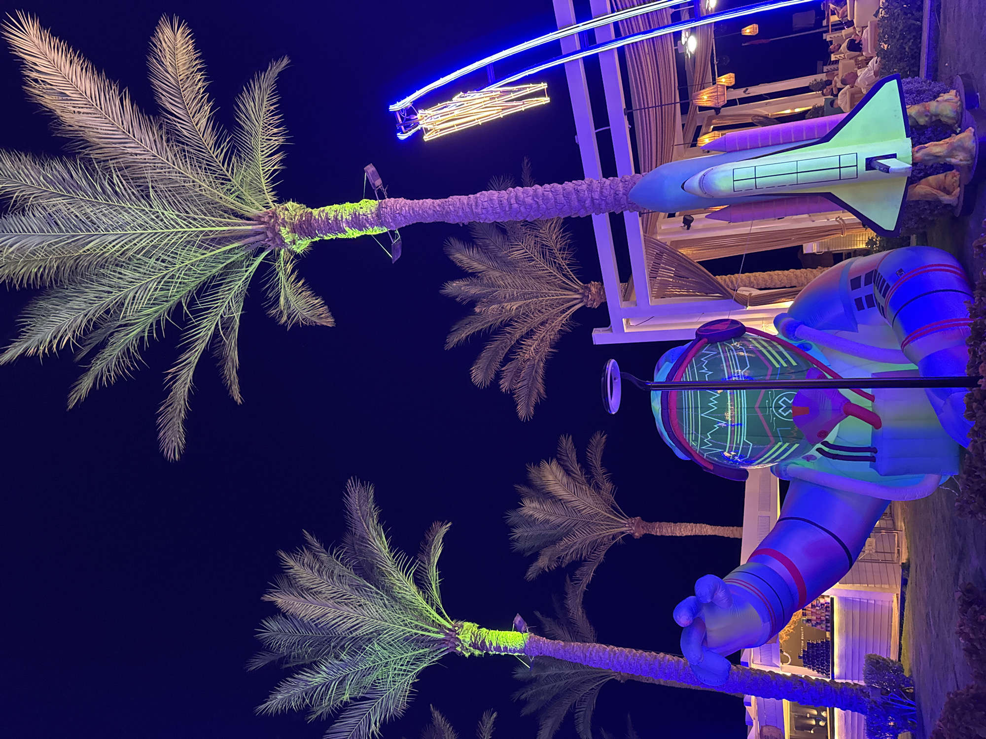 An inflatable astronaut figure stands in the foreground, with a toy rocket ship to its right, set against a nighttime scene with palm trees illuminated by colorful lights. The astronaut is predominantly blue, with green and pink accents, and its helmet displays a glowing graphic. The rocket ship is a neon yellow with pink boosters. Several palm trees are visible, their fronds illuminated with green and purple light. In the background, a white canopy structure with hanging lights and ceiling fans is set up, and there are a few people seated at tables. The scene appears to be an outdoor entertainment area, likely at a resort or hotel, given the palm trees and resort-like structures. The overall impression is one of a vibrant, nighttime celebration or attraction. The location is identified as Unknown, Egypt.