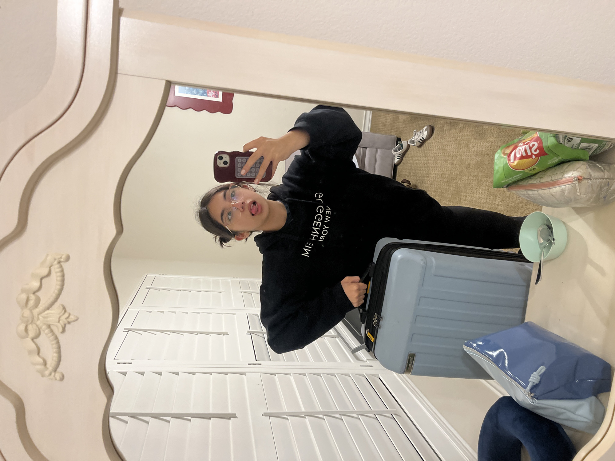 A young woman is taking a mirror selfie. She is wearing a black hoodie and glasses. Her tongue is sticking out. She is holding a phone in her right hand. Next to her is a light blue suitcase. Behind her is a white shuttered door. In the background is a bed, a gray chair, and a pair of shoes. There is also a bag of chips and a pillow on a shelf. The location cannot be determined.