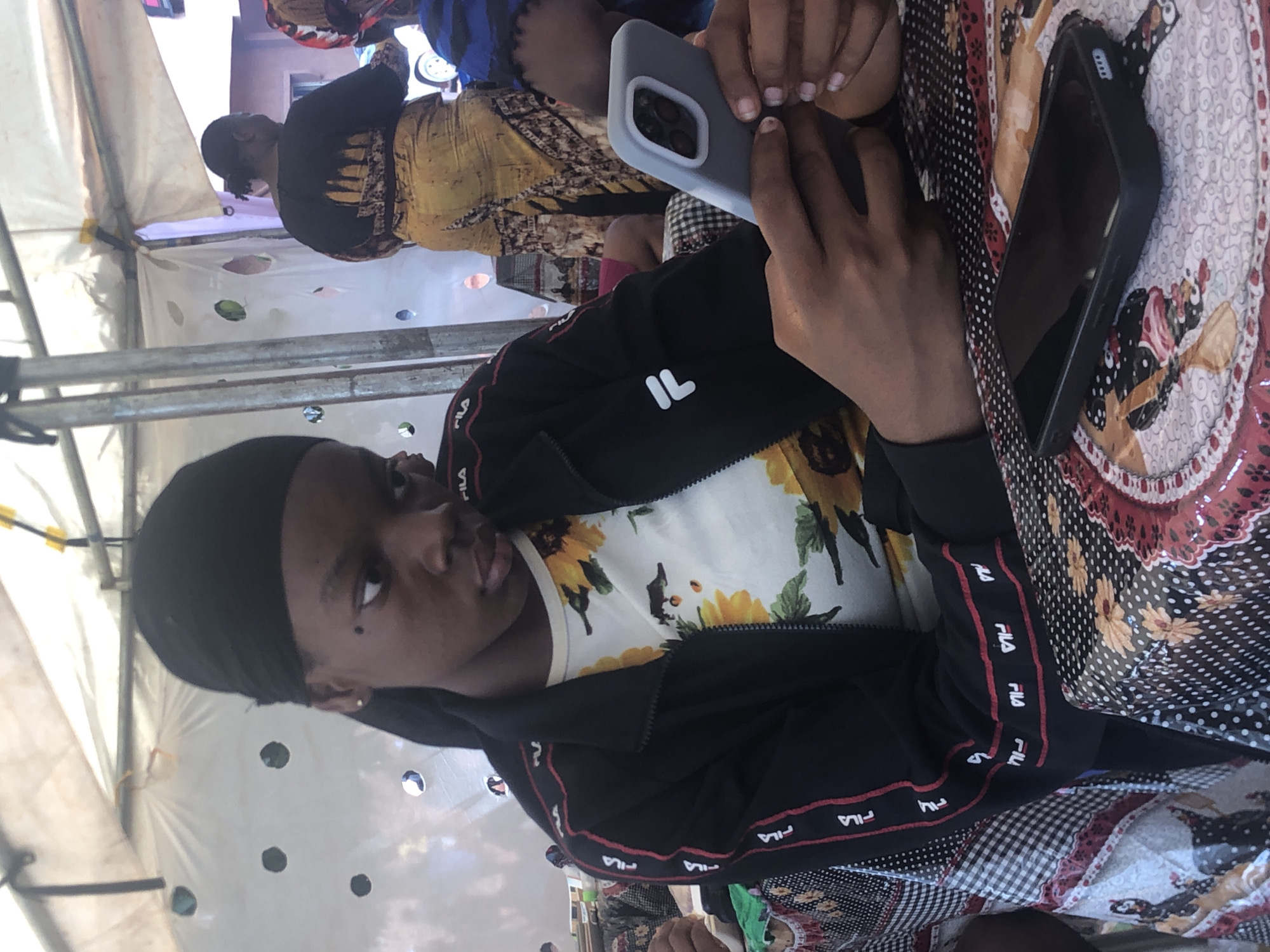 A young woman with dark hair covered by a black headscarf is holding two smartphones. She's wearing a black Fila jacket over a white sunflower-print shirt. The woman is sitting at a table covered with a patterned tablecloth. Another person, wearing a yellow and black dress, is sitting nearby. The scene appears to be outdoors, under a white tent-like structure. The location is Nteje, Nigeria.