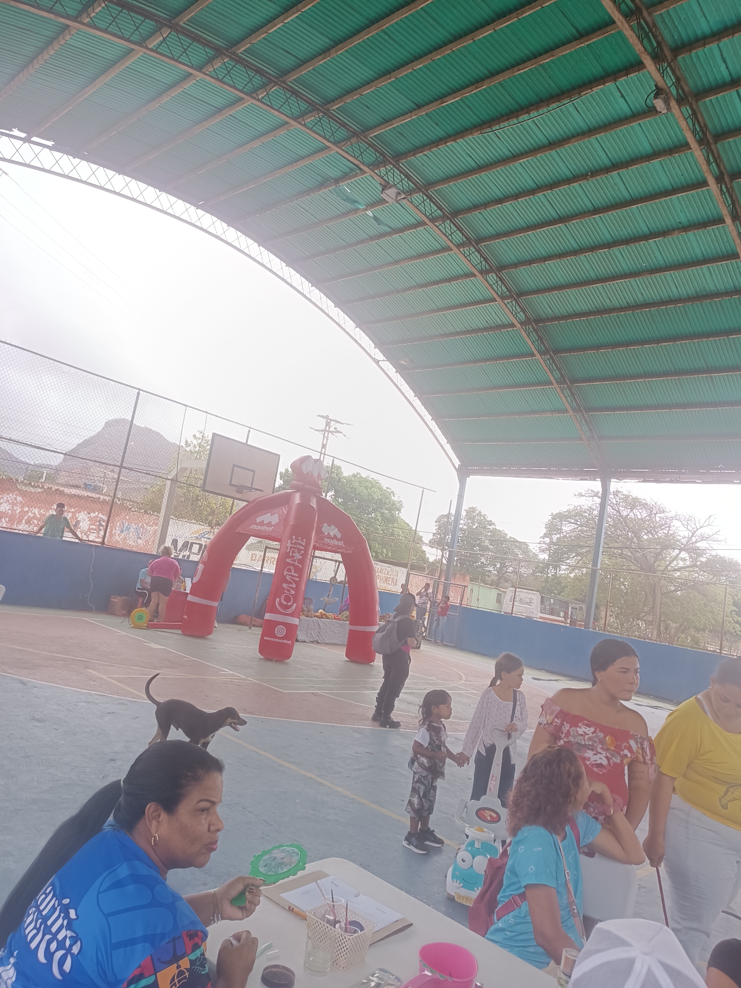 This image captures a daytime community event within a covered outdoor sports court in El Piache, Venezuela. A large, green corrugated metal roof, supported by metal trusses, provides shelter over the court. The court surface features blue and reddish-brown sections with painted lines.
In the foreground, to the left, a woman with dark hair pulled back is seated at a table. She wears a blue t-shirt with white and multi-colored patterns. She holds a green, ornate, mirror-like object in her left hand, while her right hand is positioned near a small, dark container on the table. Various craft supplies, including a white mesh container with pens/brushes, clear cups, and a pink container, are spread across the table.
A dark-coated dog, appearing to be a small to medium-sized breed, stands on the court in the midground, looking towards the right. Behind the dog, a person dressed in dark clothing and wearing a backpack is walking towards the right side of the frame.
In the midground center-right, several individuals are standing. A young girl in a dark patterned outfit is holding hands with an older girl in a white long-sleeved top and dark pants; they stand next to a white and blue toy on wheels. Behind them, a woman in a red floral off-shoulder top stands with her hands clasped, looking towards the left. To her immediate right, another woman in a yellow t-shirt and light pants is looking downwards.
In the background, a large red inflatable archway, branded with "movilnet" logos and the word "COMPARTE" (Share) written vertically, stands prominently. Below the archway, an Instagram handle "somosmovilnet" is visible. Near the base of the archway, a yellow circular object with a cord is on the ground.
The court is enclosed by a blue painted wall, behind which a chain-link fence separates the area from a natural landscape. Distant mountains or hills, sparse trees, power lines, and utility poles are visible in the background. A basketball backboard and hoop are also visible in the background, mounted on a pole near the fence. The scene is brightly lit, indicating daytime conditions, with some areas appearing overexposed.