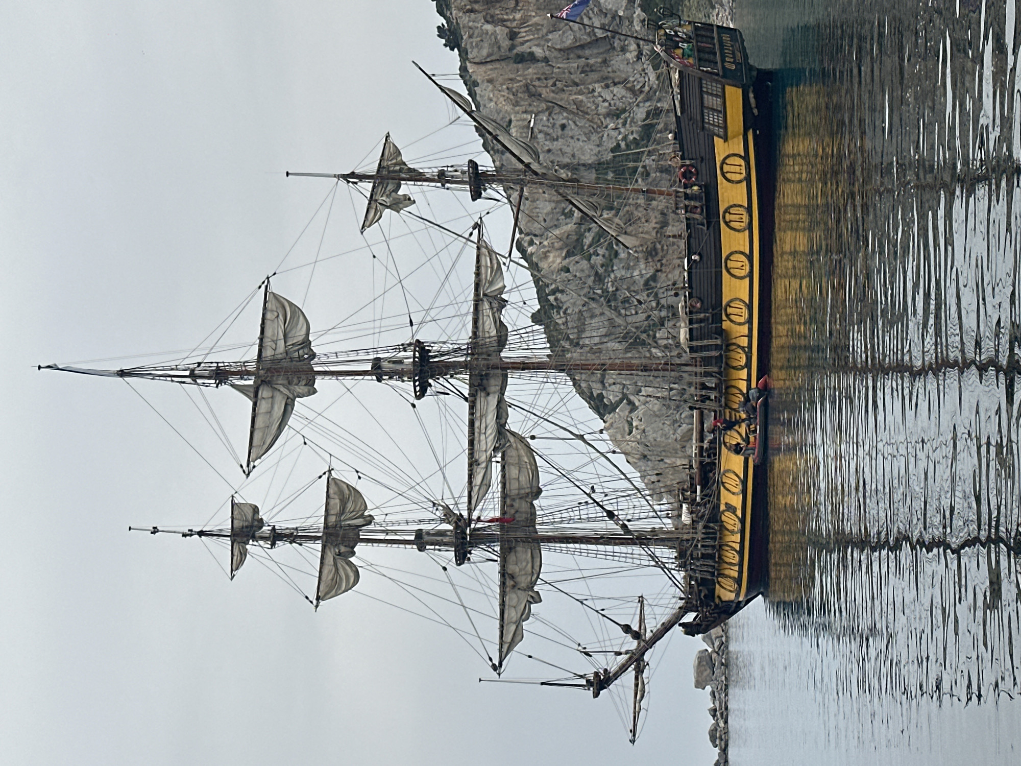 The image features a majestic multi-masted sailing ship, reminiscent of a historical tall ship or galleon, calmly floating on water against a backdrop of a rugged, light gray rocky cliff face. The ship's hull is predominantly dark, but it is accentuated by a striking yellow stripe that runs along its side. This yellow stripe is adorned with several dark, circular emblems, many of which clearly display the Roman numeral "II".
The vessel boasts at least three prominent masts, each fitted with numerous furled sails and an intricate network of ropes and rigging. Several small human figures are discernible on the ship's deck, particularly near the stern, and possibly among the rigging.
The calm water in the foreground creates distinct reflections of both the ship's hull and the textured cliff face. The sky above is uniformly overcast and light gray, casting a soft, diffused light over the entire scene. A small flag, possibly a Union Jack or similar, can be seen flying near the ship's stern, atop one of its masts. Faintly visible on the ship's stern, above a row of windows, is the text "MARTELLOO".
The scene suggests the ship is either anchored or docked close to the shore in a bay or harbor. The peaceful coastal setting, with its dramatic cliff, aligns with locations such as Datça, Turkey, as provided in the context. The ship appears to be either a well-maintained historical vessel or a meticulously crafted replica.