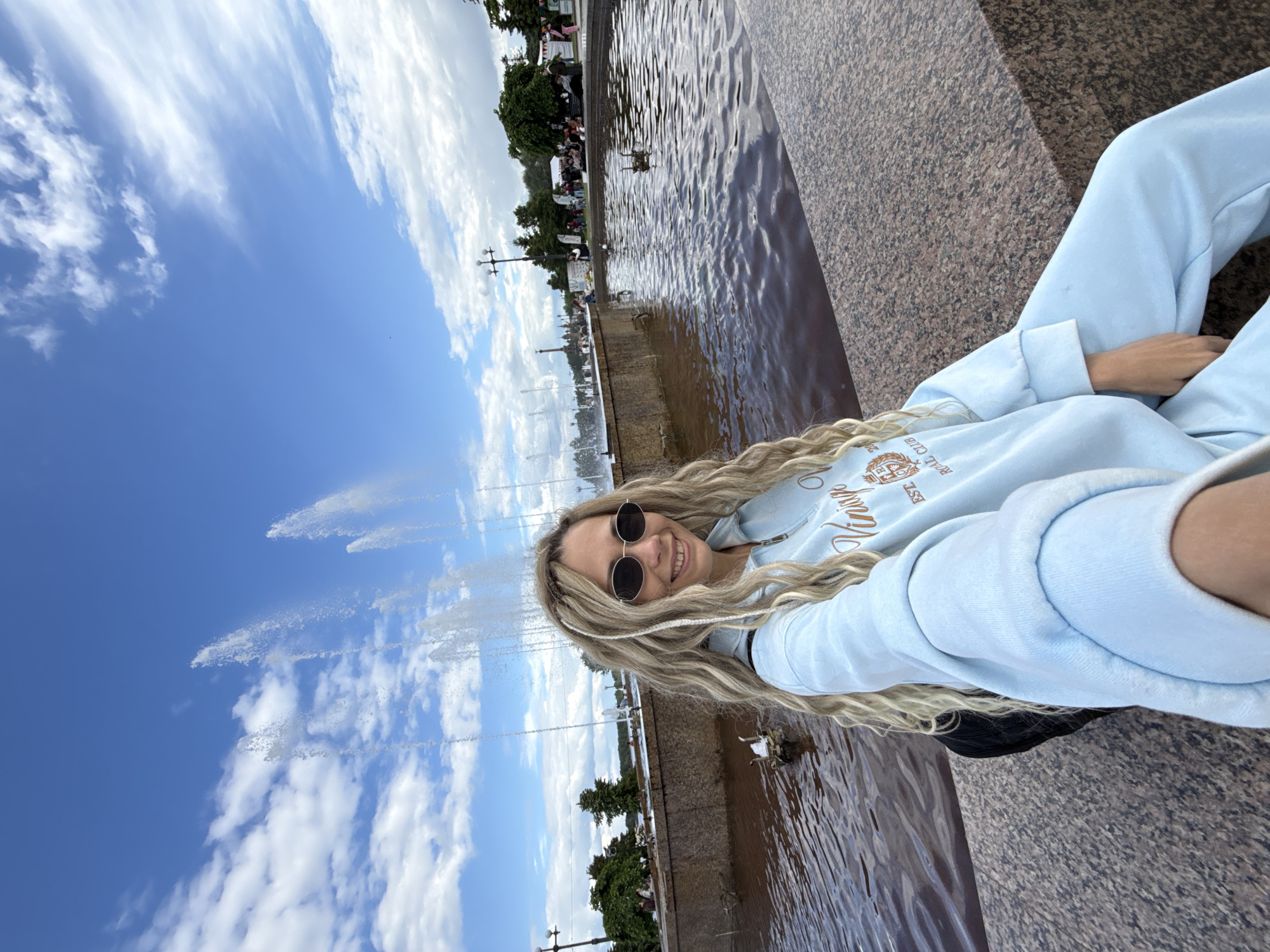 A woman with blonde hair and sunglasses is sitting next to a fountain in the city of Rusanovo, Russia. She is wearing a light blue sweatshirt and sweatpants. The fountain has multiple jets of water spraying into the air. The sky is blue with white clouds. There are trees and buildings in the background. People are sitting on benches in the background. A duck is swimming in the fountain.