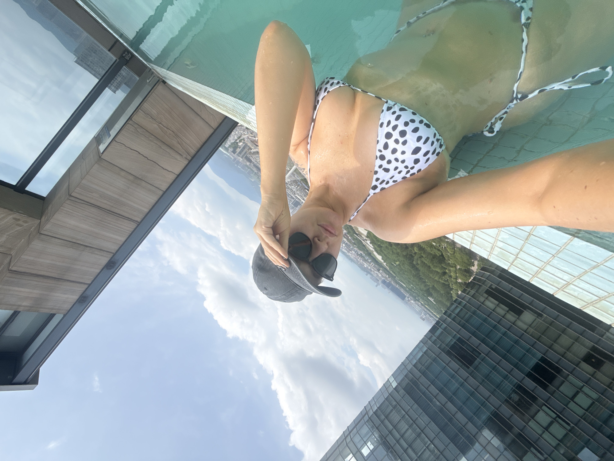 A woman in a black and white polka dot bikini and a dark baseball cap poses for a selfie in an infinity pool. She is wearing sunglasses and resting her hand on the brim of her cap. The pool overlooks a cityscape with tall buildings, including a glass-fronted skyscraper, and a lush green treeline. The sky is blue with scattered white clouds. The overall scene suggests a luxury rooftop setting in a city.