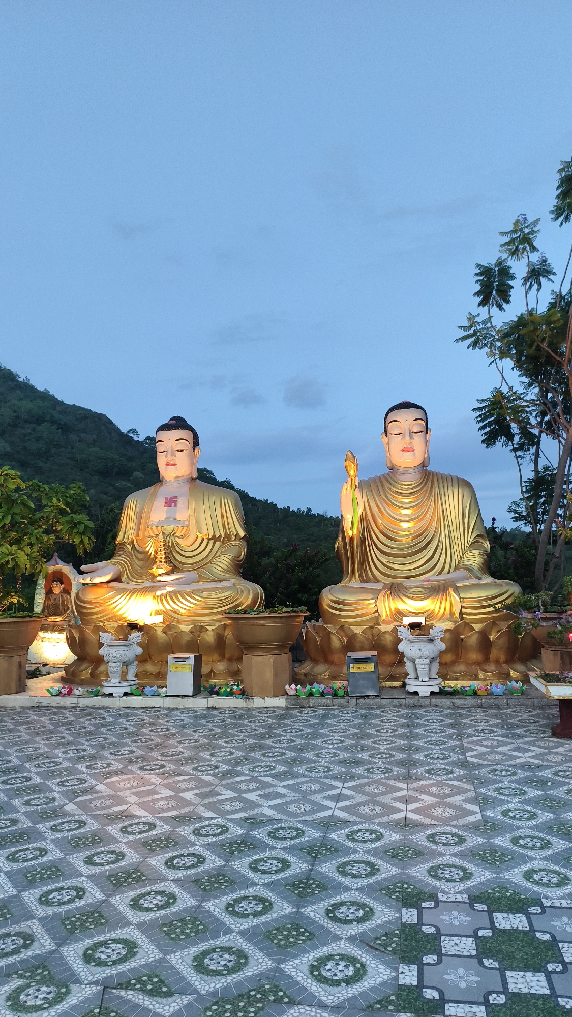 Two large, golden Buddha statues are seated on lotus bases, illuminated by artificial lights against a twilight sky and a backdrop of lush green hills. The statue on the left has a swastika symbol on its chest. The statue on the right holds a lotus flower. In front of the statues are smaller offerings and decorative elements, including potted plants and ceramic figures. The foreground features a patterned tiled floor with geometric designs. The scene is set in a tranquil outdoor environment, likely a temple or spiritual site, suggesting a location in Bac Nha Trang Ward, Vietnam, a region known for its Buddhist pagodas and natural beauty.