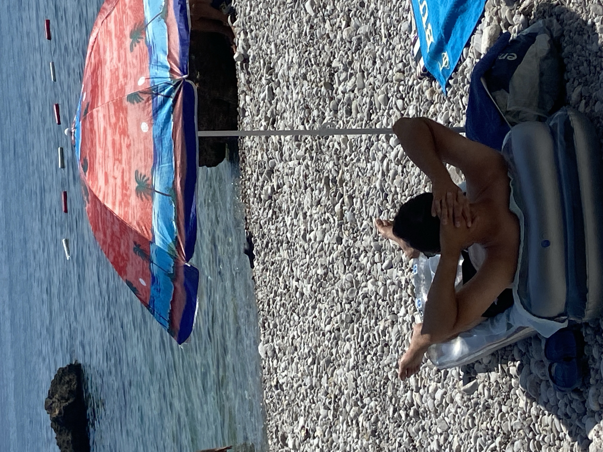 The image depicts a person relaxing on a pebble beach in Prijevor, Montenegro, on a sunny day. The person is lying on an inflatable mat, facing away from the camera, with their arms behind their head. They are wearing dark swimwear. Beside them, a colorful beach umbrella with a palm tree design is open, casting a shadow on the pebbles. A blue towel is visible in the background, along with a dark blue bag. The sea is calm and blue, with a few small rocks visible in the water. The sun appears to be high in the sky, suggesting it is midday. The overall mood is one of relaxation and leisure. Visible text on the blue towel is partially obscured.