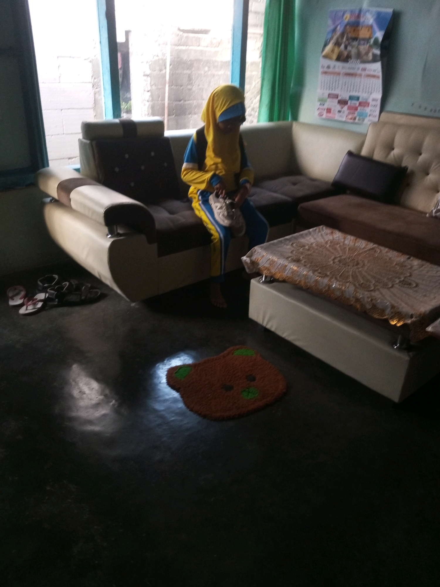 A person wearing a yellow hijab and blue and yellow athletic wear sits on a sectional couch in a room. The couch is made of light-colored faux leather with dark brown cushions and a dark brown accent pillow. To the right of the person, a low coffee table is covered with a patterned tablecloth. In the foreground, a brown rug shaped like a bear's face lies on the dark floor. Several pairs of sandals are scattered near the couch. A calendar hangs on the wall behind the couch, and a window with sheer green curtains is visible to the left. The room appears to be indoors, possibly a living room.
