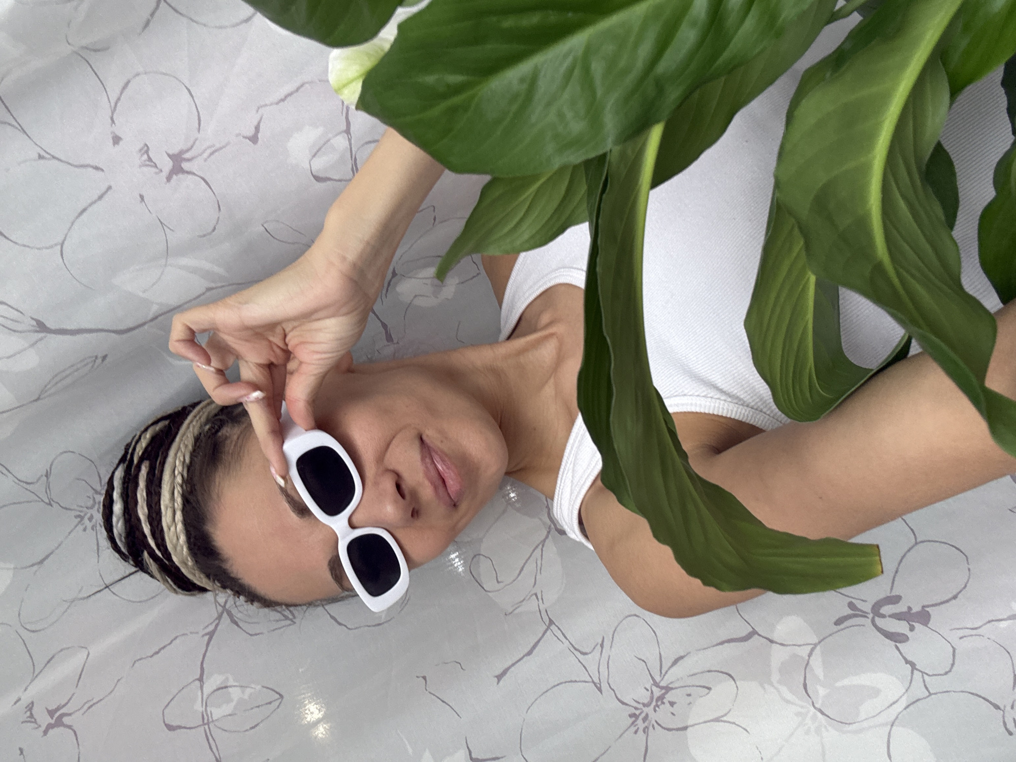 A woman is lying on a surface patterned with stylized floral designs. She wears a white tank top and white-framed sunglasses. Her hair is styled in black and blonde braids. The woman's hand is near her sunglasses. Large, green leaves of a plant partially obscure her body. There is no clear indication that this image was taken in Rusanovo, Russia.