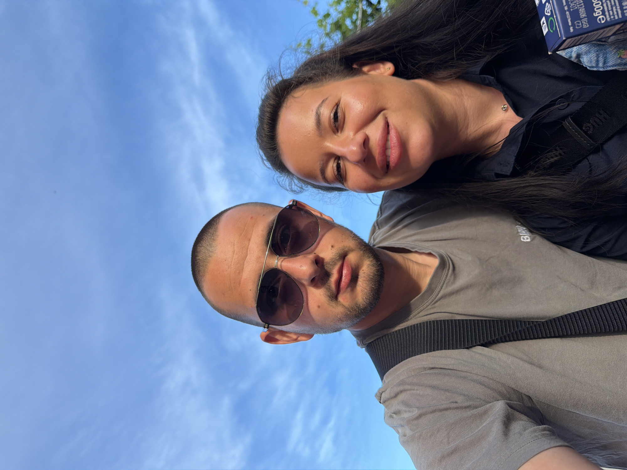 A selfie captured from a low angle shows a man and a woman outdoors against a bright blue sky with wispy clouds. The man, positioned on the left, wears a grey t-shirt and sunglasses, with a dark strap visible across his chest. The woman, on the right, has long dark hair and wears a dark collared shirt. She is smiling and looking towards the camera. A blue cardboard box is partially visible in her lower right corner. The overall setting suggests a casual outdoor environment.