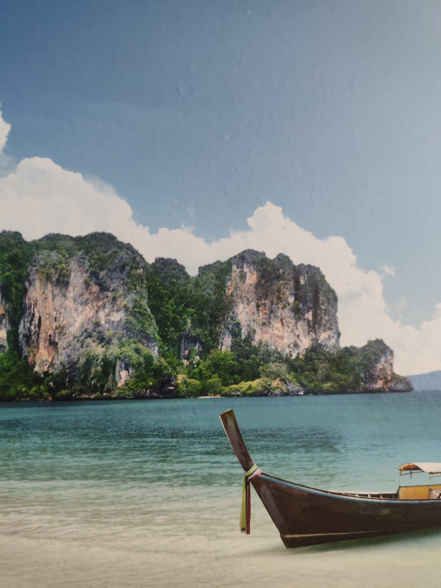 The image displays a tropical coastal scene in Unknown, Unknown, under a light blue sky with scattered white cumulus clouds. In the mid-ground, a series of large, imposing limestone karsts, heavily covered in lush green foliage, rise steeply from the water. Their exposed rock faces exhibit a variety of textures and colors, including grays, browns, and reddish tones. The ocean in front of the islands is a deep turquoise, transitioning to a lighter aquamarine color with visible ripples as it extends towards the foreground. A light-colored sandy beach occupies the bottom third of the frame. In the right foreground, a dark brown longtail boat rests partially on the sand, its high, pointed bow angled towards the left. A decorative fabric ribbon, striped with yellow, green, and red, is tied to the boat's prow. The stern of the boat features a light-colored canopy over an enclosed compartment, with yellow accents visible beneath. No people are present in the scene.