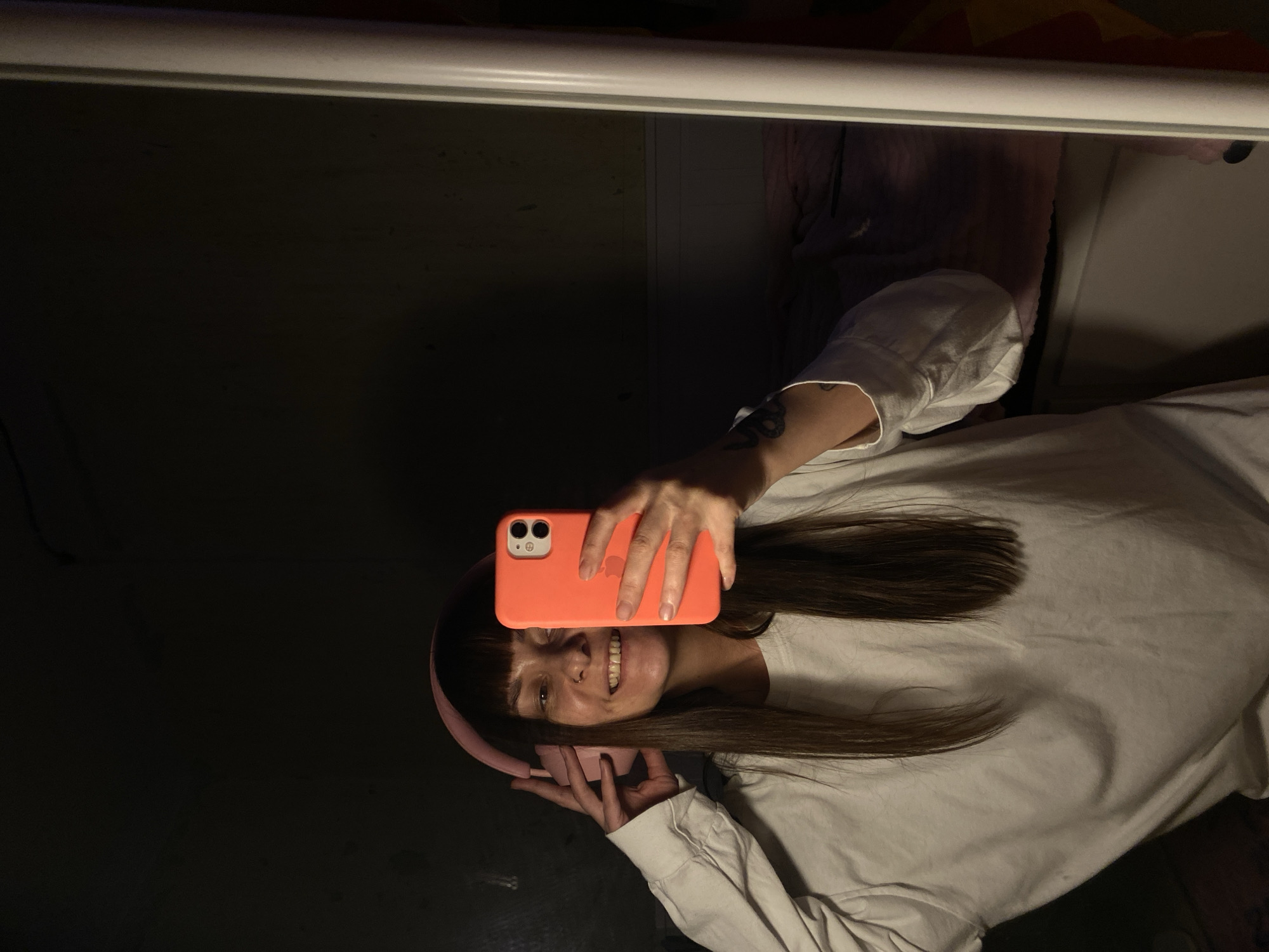 A woman with brown hair and bangs is holding an orange smartphone and smiling. She is wearing a white long-sleeved shirt and pink headphones around her neck. The background is partially dark and partially lit, revealing a white surface and some indistinct shapes. There is no way to determine from this image whether the location is Votkinsk, Russia, or any other city.