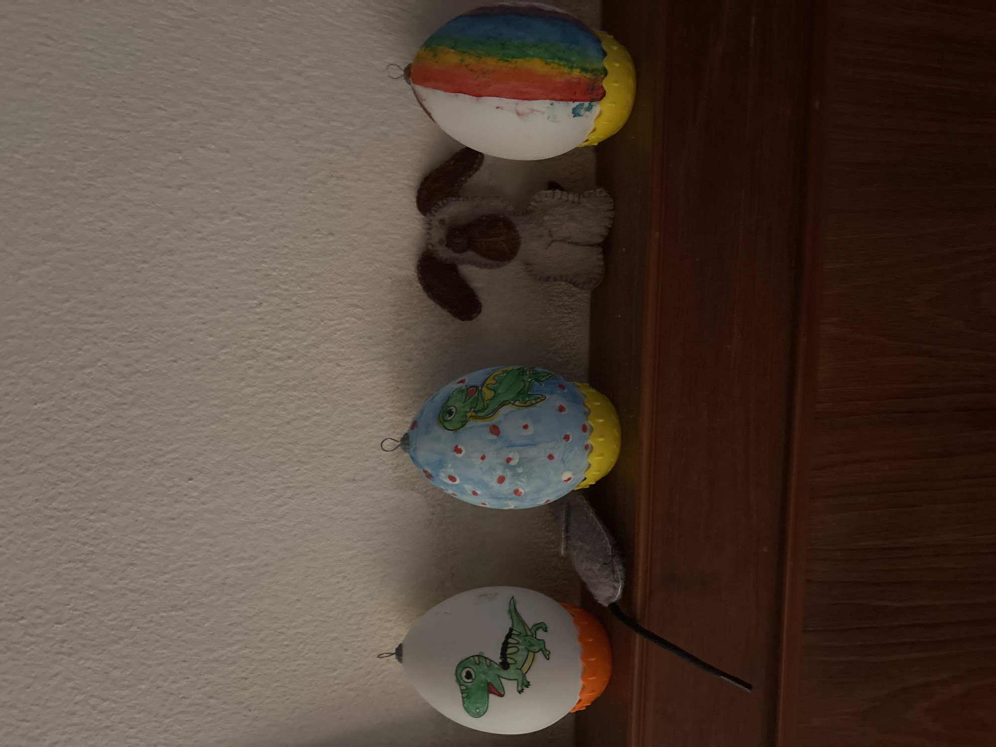 The image shows three painted, egg-shaped objects hanging vertically along a wall. The top object is white with a rainbow pattern. Below it is a gray stuffed toy resembling an animal. The second egg-shaped object is blue with a green dinosaur pattern. Below it is a gray, fuzzy object and a black wire. The third egg-shaped object is white with a green dinosaur pattern and an orange bottom. The wall is a light color, and a dark wood surface is visible on the right side of the image. There is no evidence to support the location in บ้านบางรักษ์, Thailand.