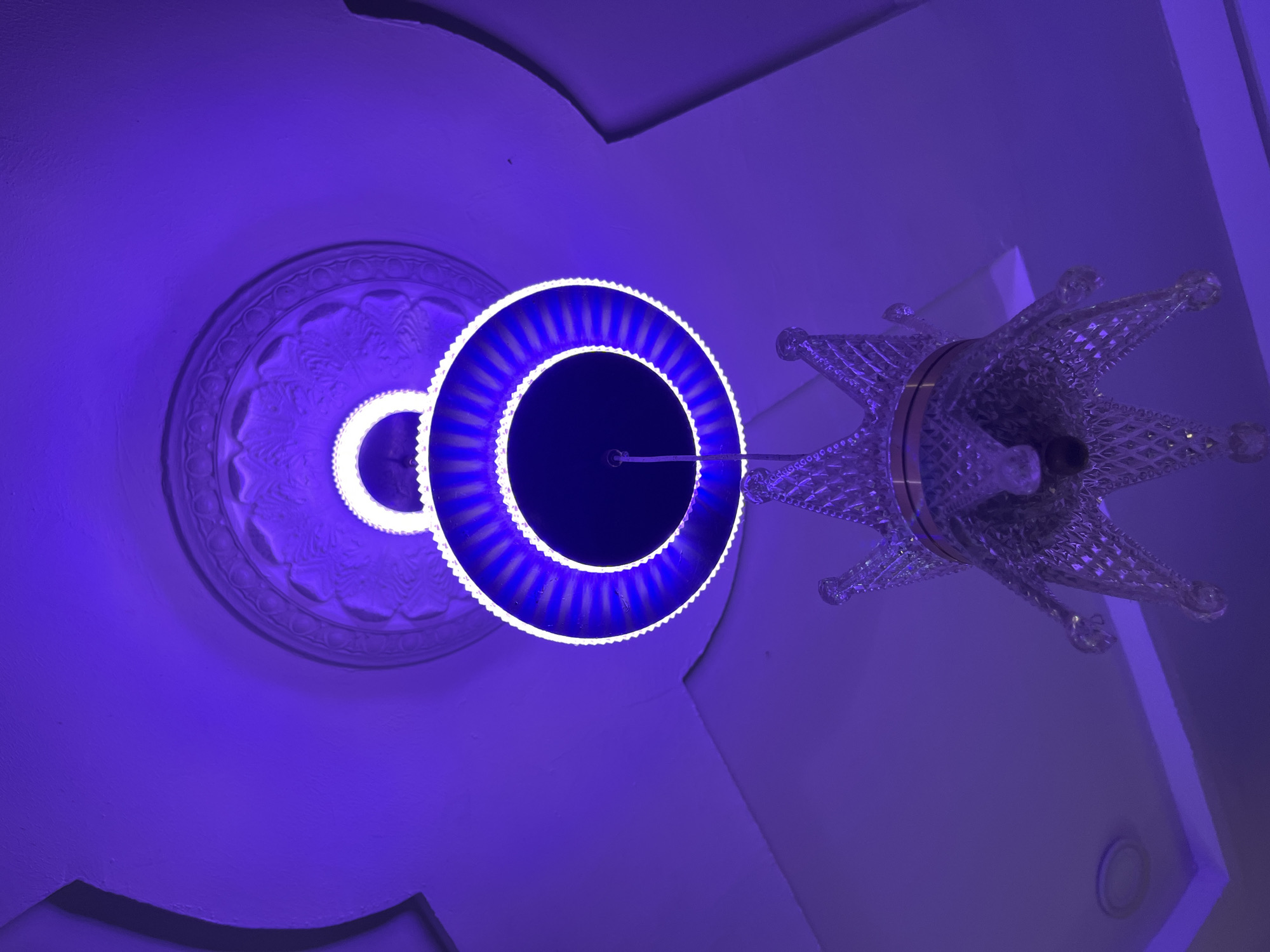 The image captures a close-up, low-angle view of a ceiling with ornate decorative molding and a modern chandelier. The lighting is predominantly a vibrant purple hue, suggesting artificial illumination. The chandelier itself is a striking object, featuring a circular LED light fixture with blue and white accents, surrounding a dark central area. To the right of the LED ring, a portion of a decorative crown-like structure made of clear, faceted material is visible. This crown element appears to be part of the chandelier, adding a touch of regal ornamentation. In the background, a section of the ceiling shows detailed plasterwork in a circular pattern, also bathed in the purple light. The overall scene creates an atmospheric and somewhat dramatic effect due to the intense color. The setting appears to be indoors, likely a room with a decorative ceiling. No people are visible, and the focus is entirely on the architectural details and the illuminated chandelier. The location context provided is Takau, Nigeria, but there are no specific visual cues within the image to confirm this. The time of day and weather are indeterminate due to the indoor setting and artificial lighting. No text is visible in the image.