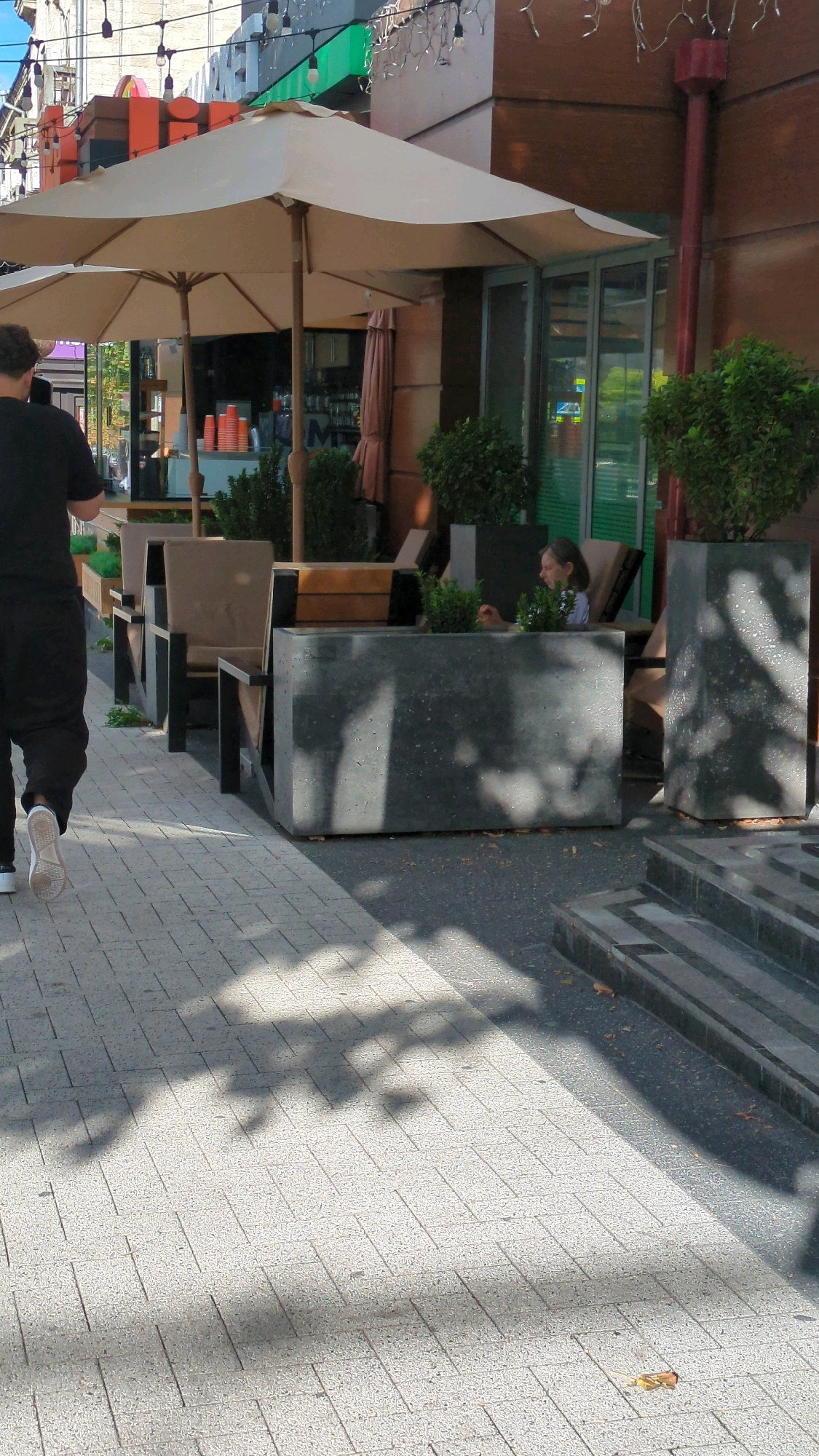 The image depicts an outdoor seating area of a cafe or restaurant on a paved walkway in Chișinău, Moldova. A person dressed in black pants and a black shirt with white sneakers is walking away from the camera towards the left side of the frame. In the background, there are outdoor tables with chairs under large beige umbrellas, and planters with green bushes. A young girl with light brown hair wearing a light blue top is seated at one of the tables, looking to her right. The building facade behind the seating area is made of brown material with glass doors and windows. Overhead string lights are visible, suggesting an evening or a dimly lit area. The ground is paved with light-colored rectangular tiles, and shadows of trees or awnings are cast across the walkway and seating area.